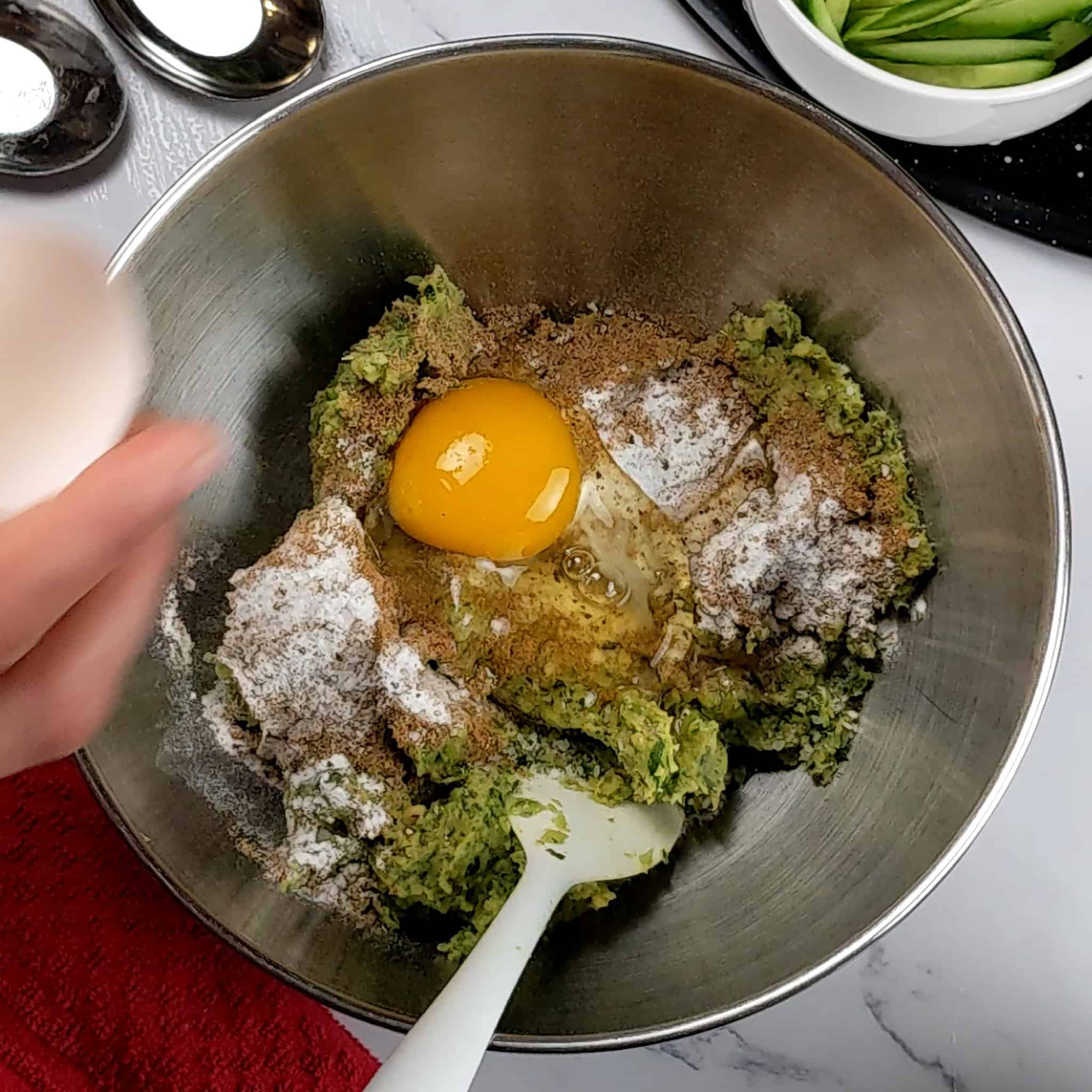 a stainless steel mixing bowl with the blended chickpea herb mixture with a raw egg, and baking soda with a spatula inside.
