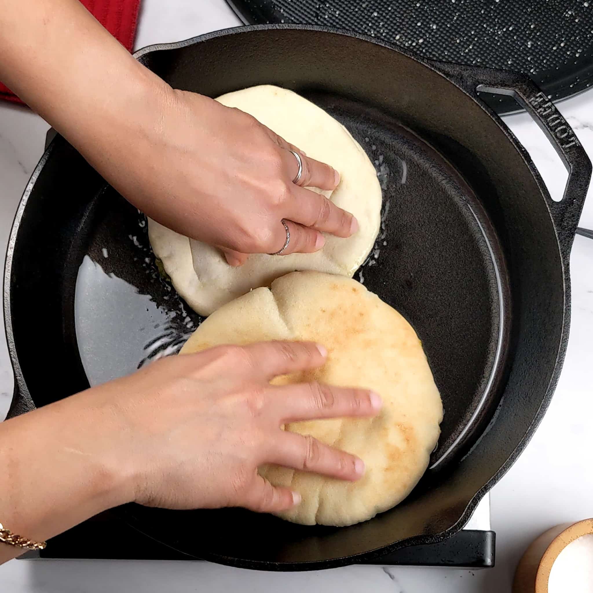 two pitas being pressed down by two hands in an oiled cast iron skillet.