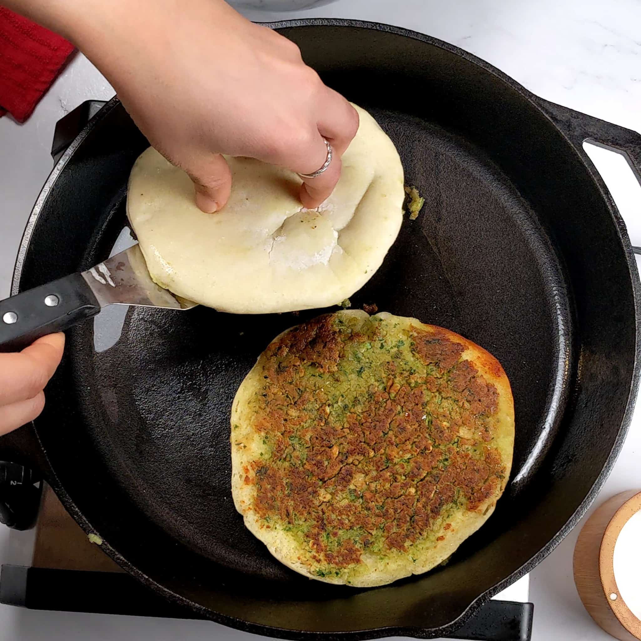 two falafel pitas in a cast iron skillet, one is falafel side up golden brown while the other is in the middle of being flipped with an offset spatula.