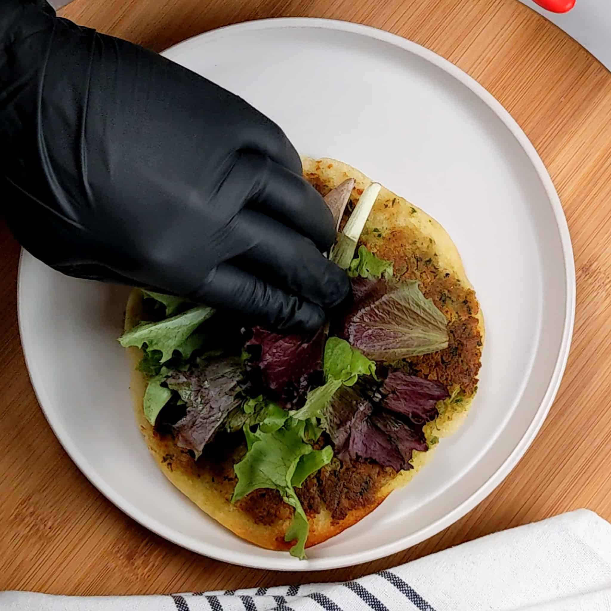 fresh mixed greens being added by a gloved hand to the top of the pita's falafel side on a round plate that is on a lazy susan next to a kitchen towel.