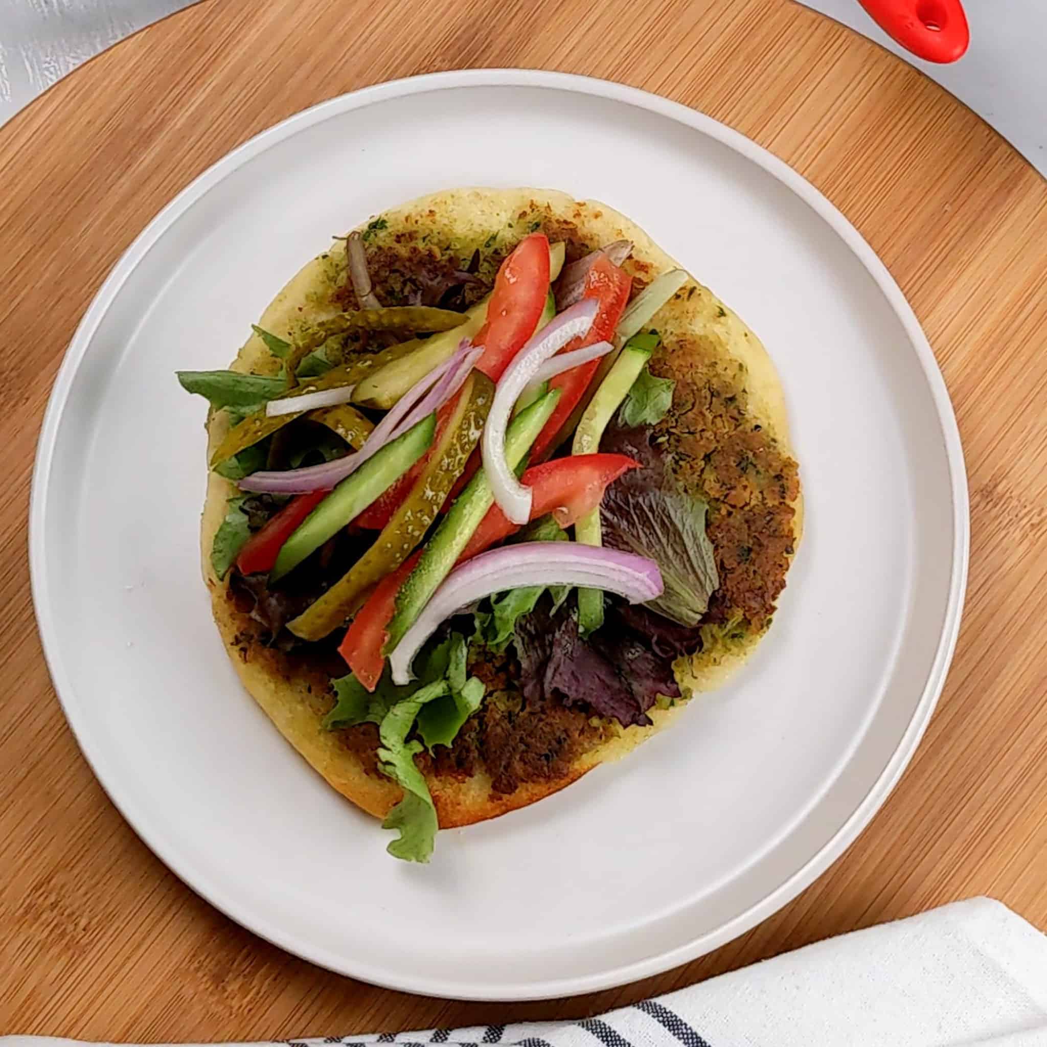 vegetable strips of red onion, tomato, pickles and cucumber added to the top of fresh mixed greens on the pita's falafel side on a round plate that is on a lazy susan next to a kitchen towel.