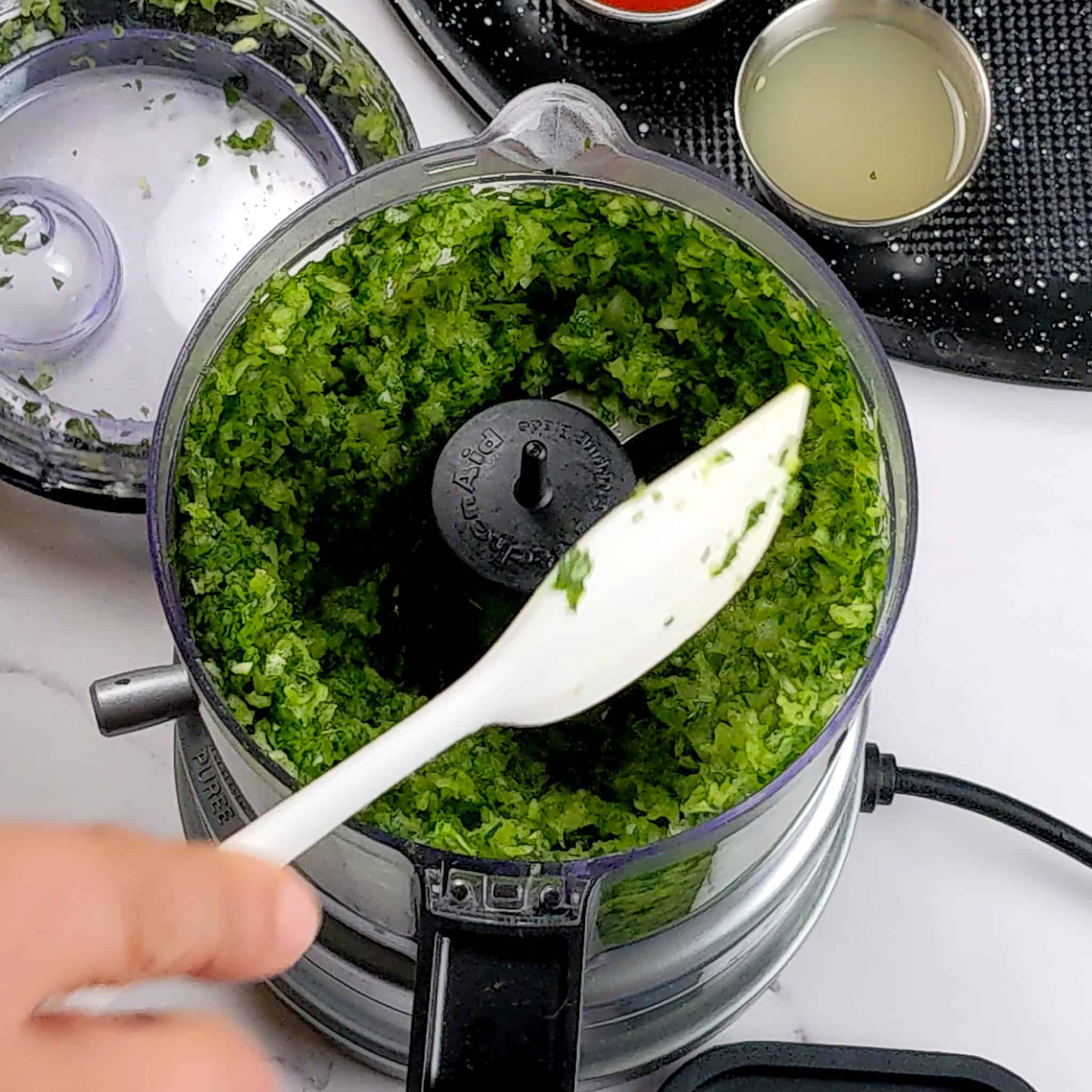 blended herbs, onion and garlic in a food processor being scrapped down with a small silicone spatula.