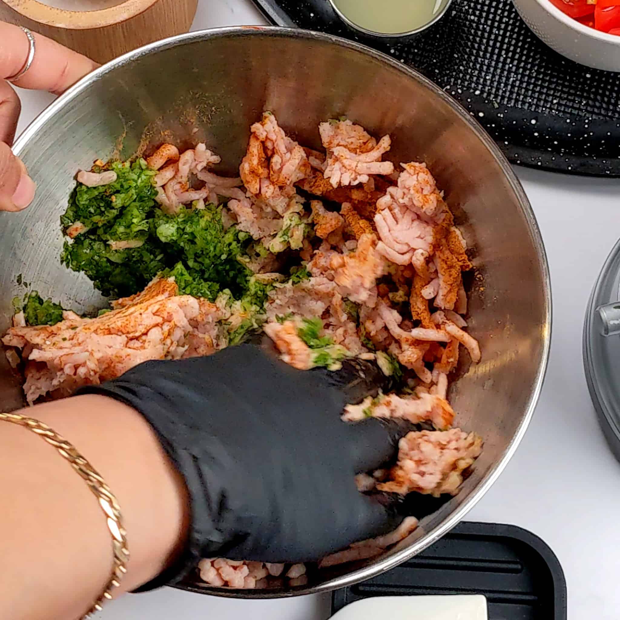 ground meat with aromatic paste and seasoning in a stainless steel bowl being mixed with a gloved hand.