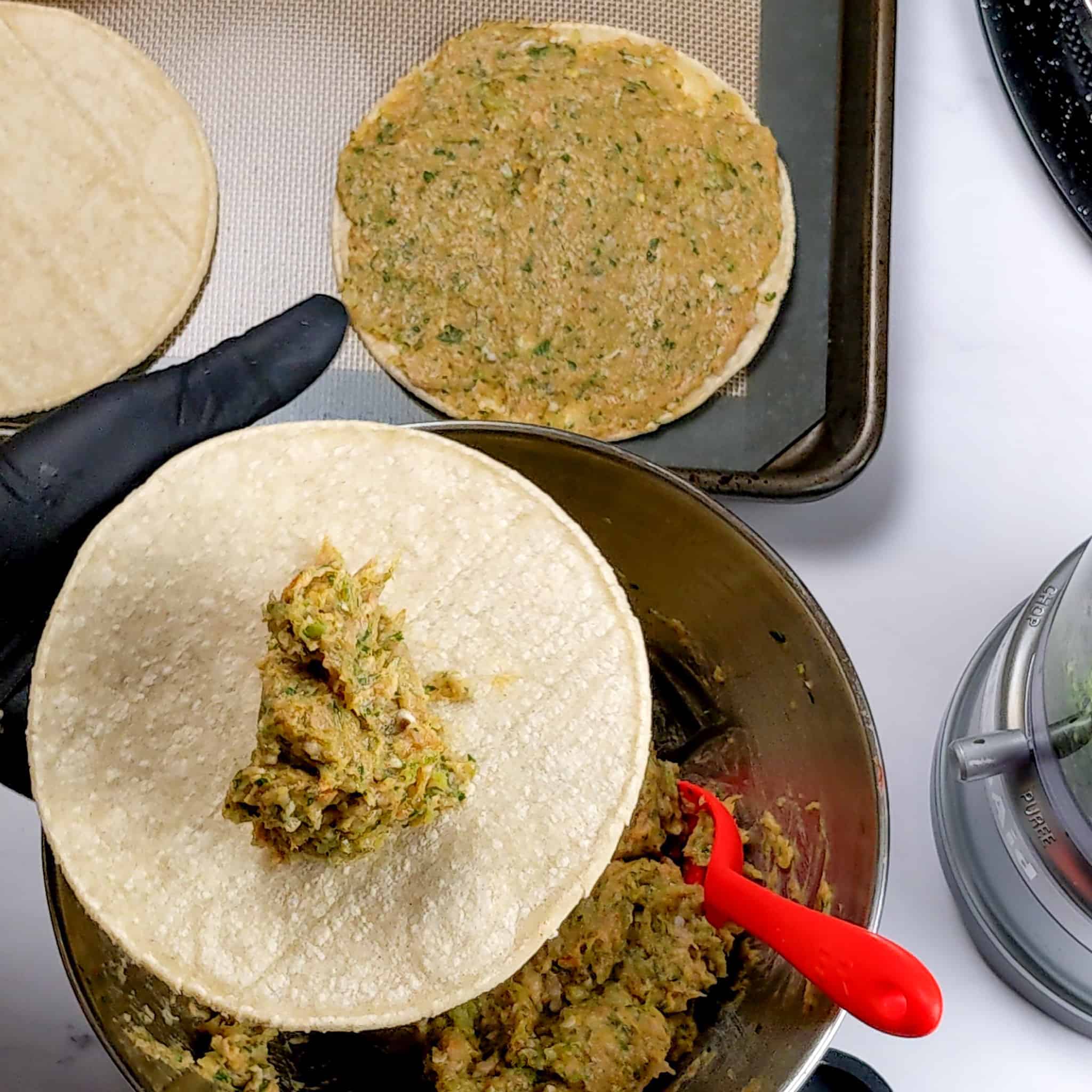 a spoonful of chicken taco meat on a corn tortilla being held in a gloved hand over the bowl of taco meat with a silicon spoon resting on it.