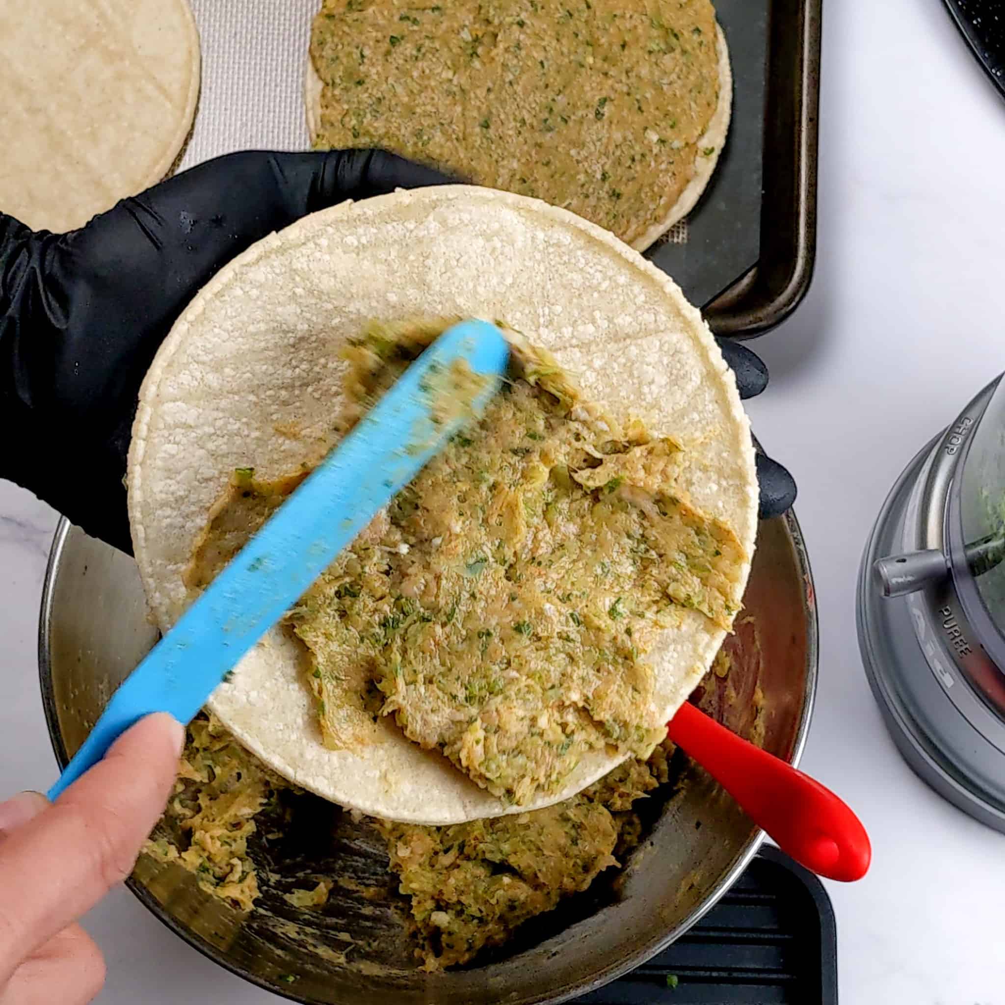 a long silicone spatula is being used to spread out the taco meat mixture onto a corn tortilla being held in a gloved hand.
