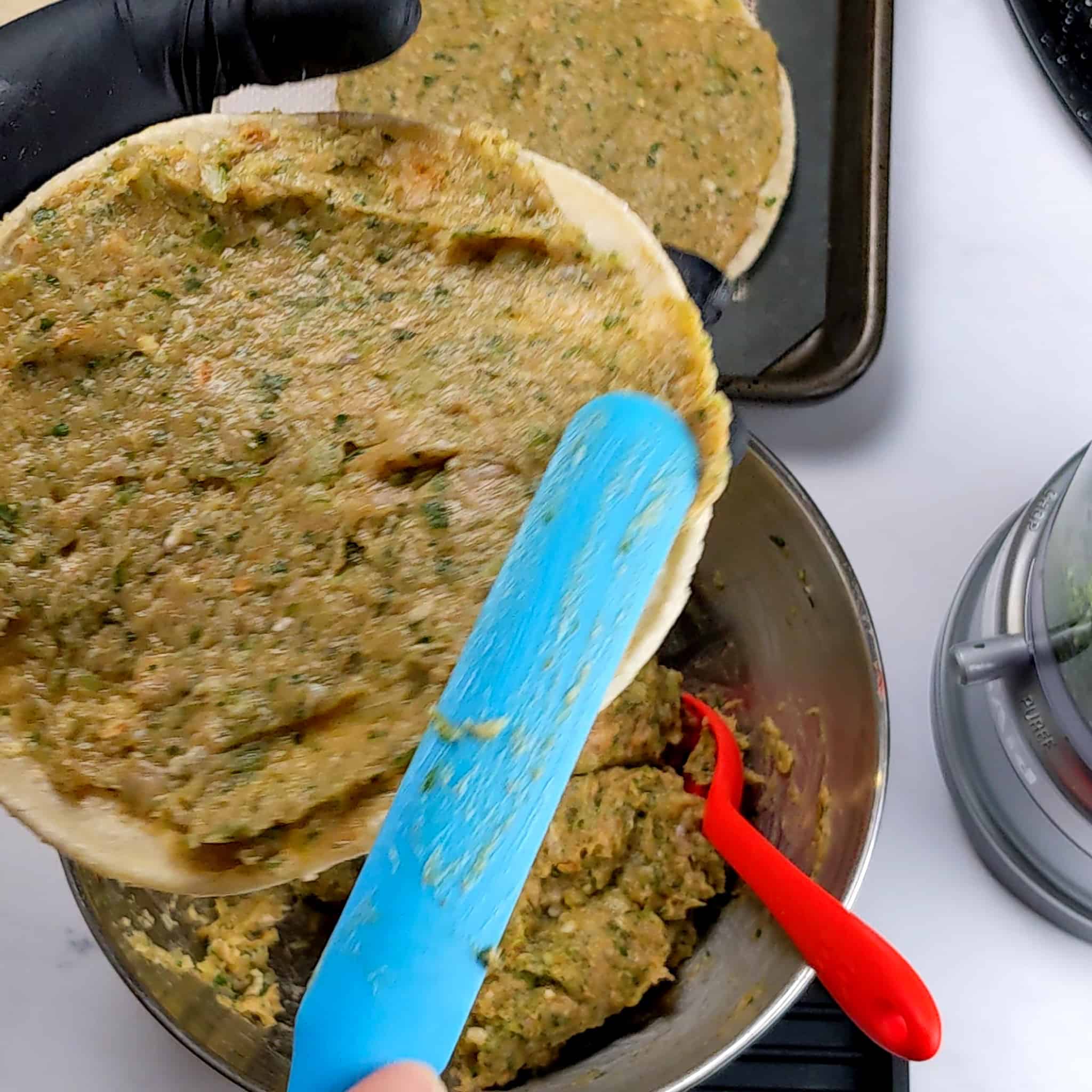 a long silicone spatula is being used to spread out the taco meat mixture to the edges of a corn tortilla being held in a gloved hand.