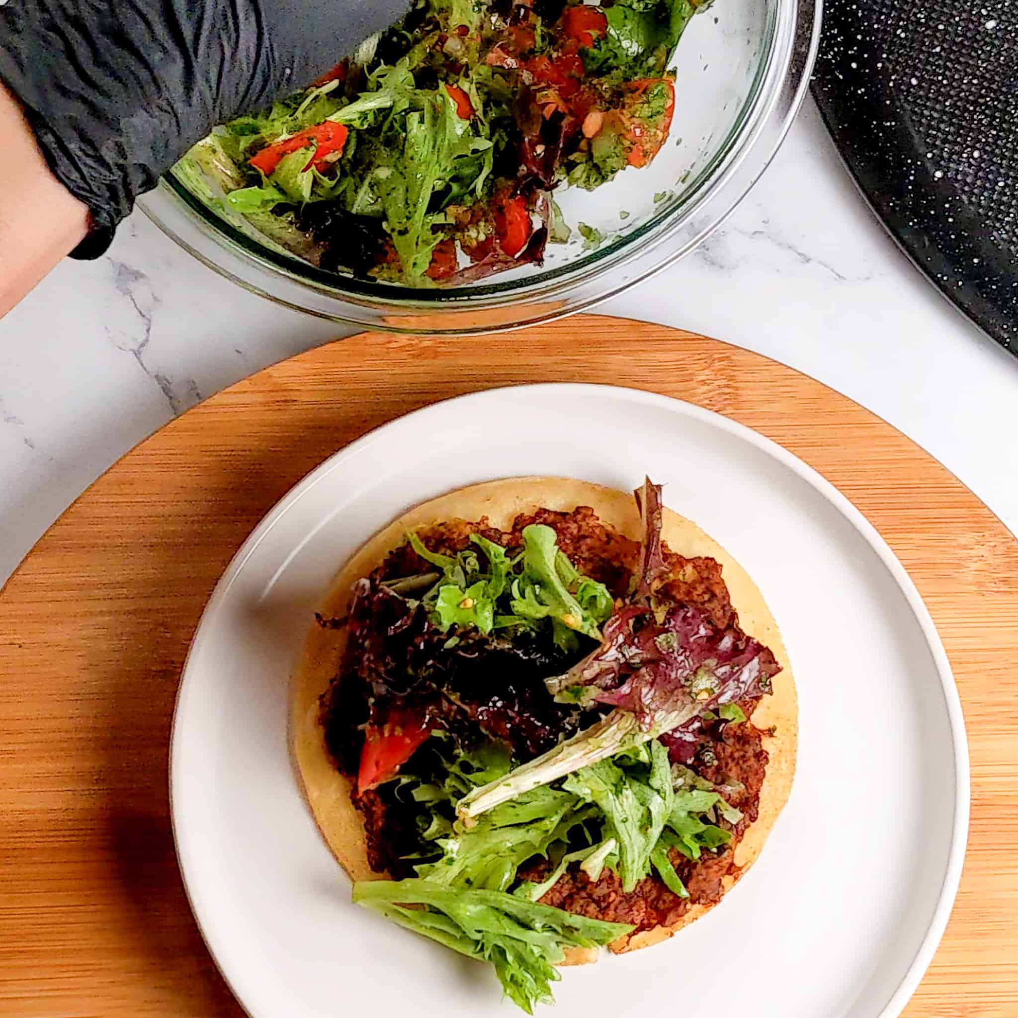 chicken taco with tomato mixed salad being added to the top on a round plate on a wooden lazy susan