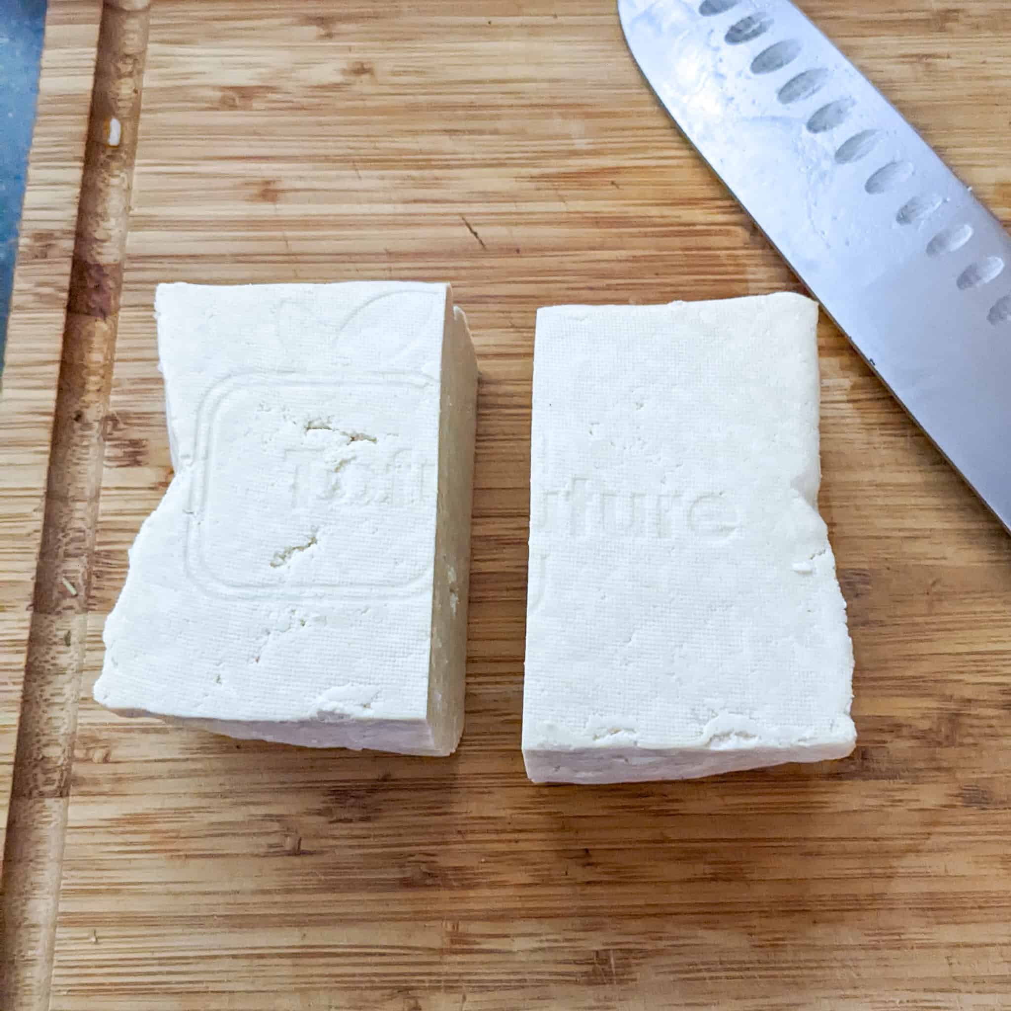 pressed tofu on a wooden cutting board cut into half displayed with a santoku knife