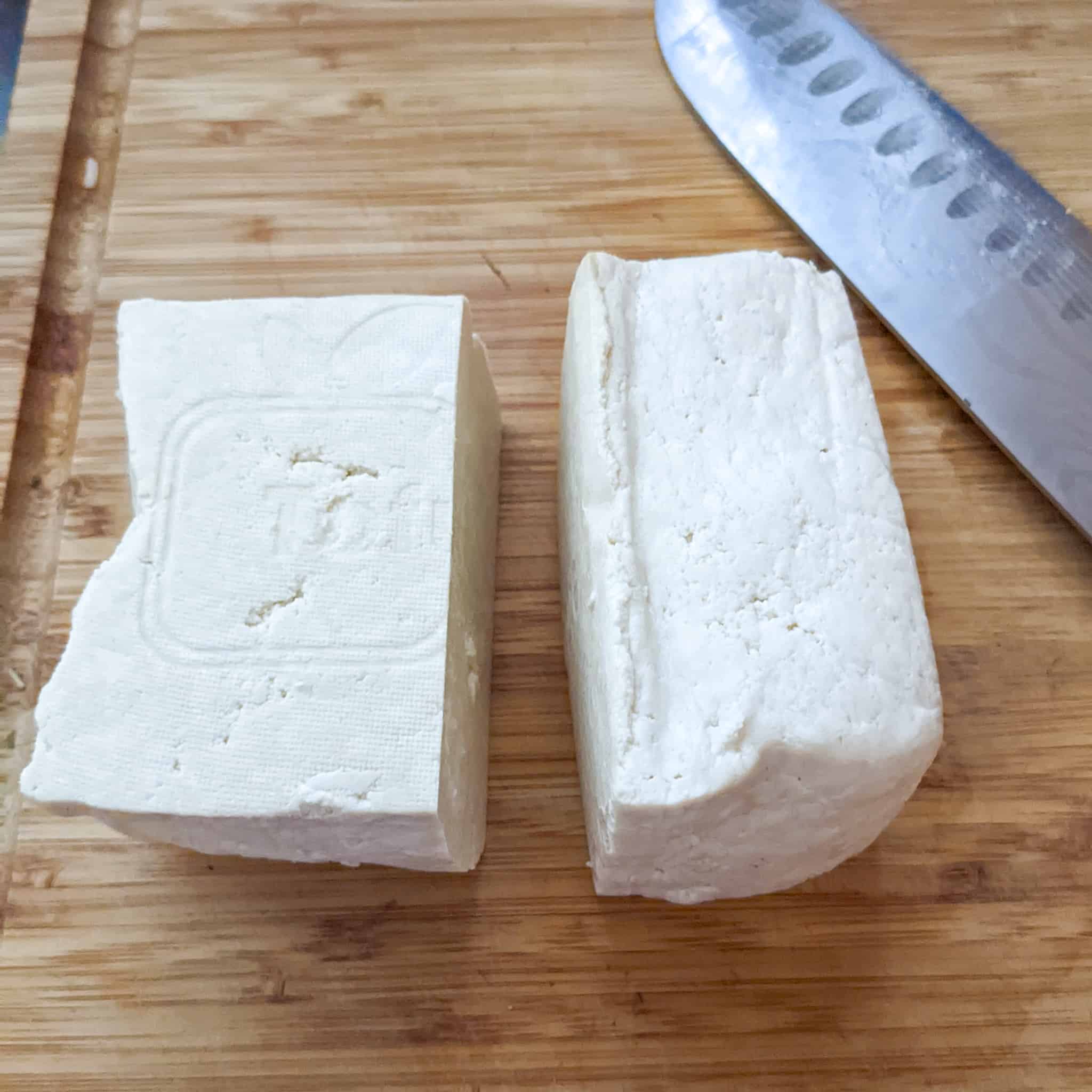 pressed tofu on a wooden cutting board cut into half with one piece placed on it's side displayed with a santoku knife