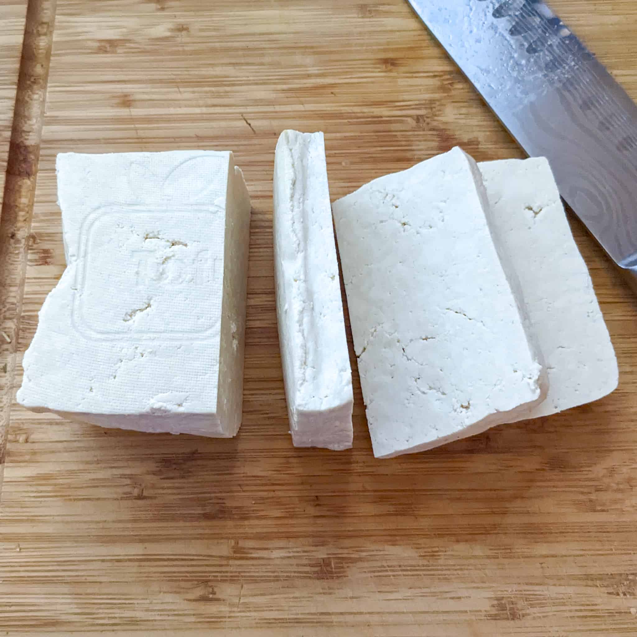 pressed tofu on a wooden cutting board cut into halves with one piece cut into three slices displayed next to a santoku knife