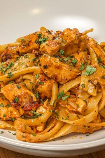 Simple Spicy Garlic Chili Sauce Chicken Pasta in a wide rim bowl with a kitchen towel and lodge enameled cast iron dutch oven with the remaining pasta dish in the background.