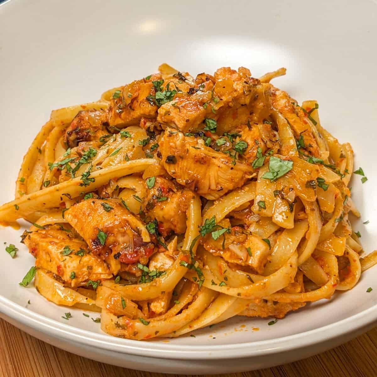 Simple Spicy Garlic Chili Sauce Chicken Pasta in a wide rim bowl with a kitchen towel and lodge enameled cast iron dutch oven with the remaining pasta dish in the background.