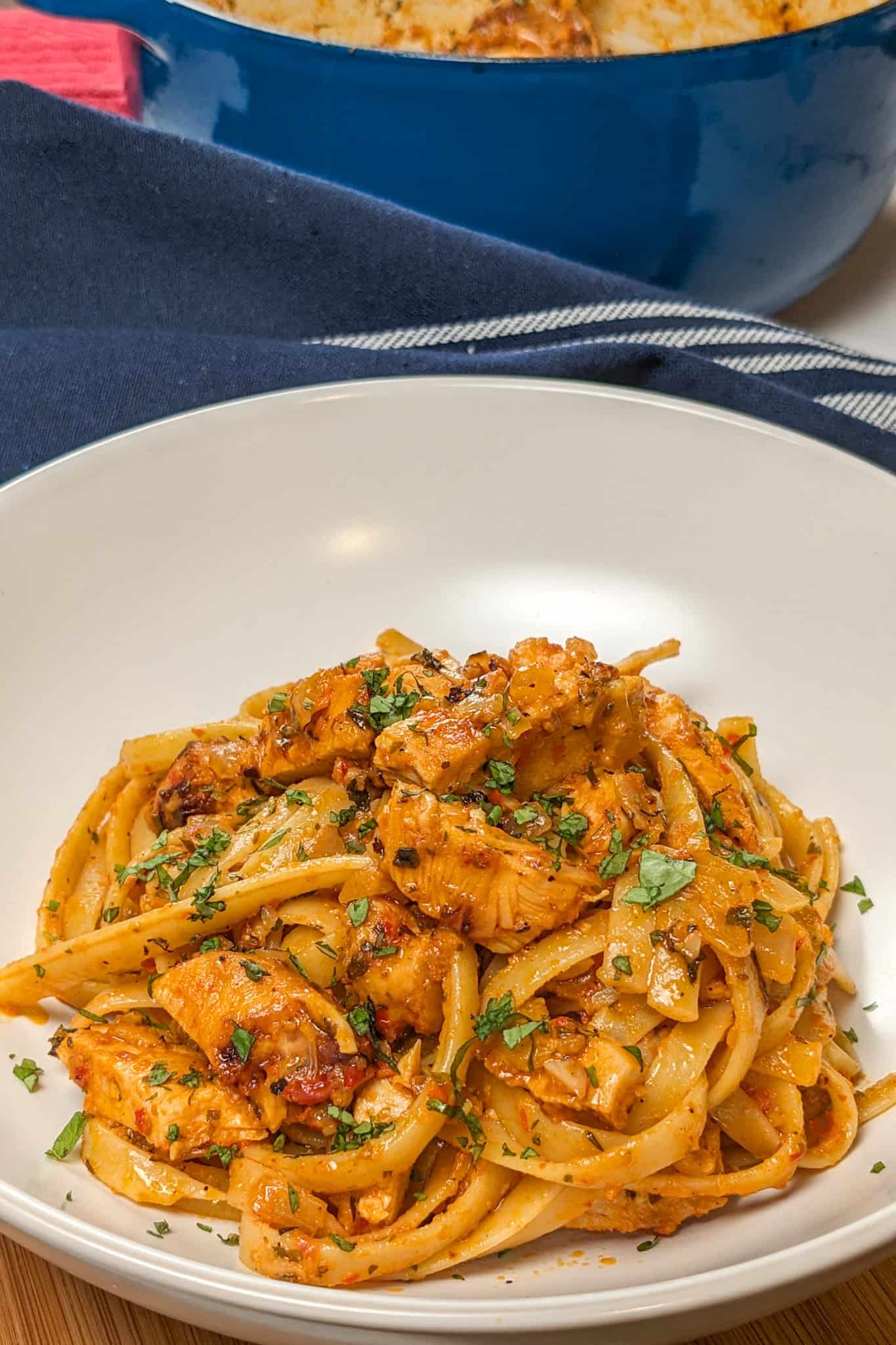 Simple Spicy Garlic Chili Sauce Chicken Pasta in a wide rim bowl with a kitchen towel and lodge enameled cast iron dutch oven with the remaining pasta dish in the background.