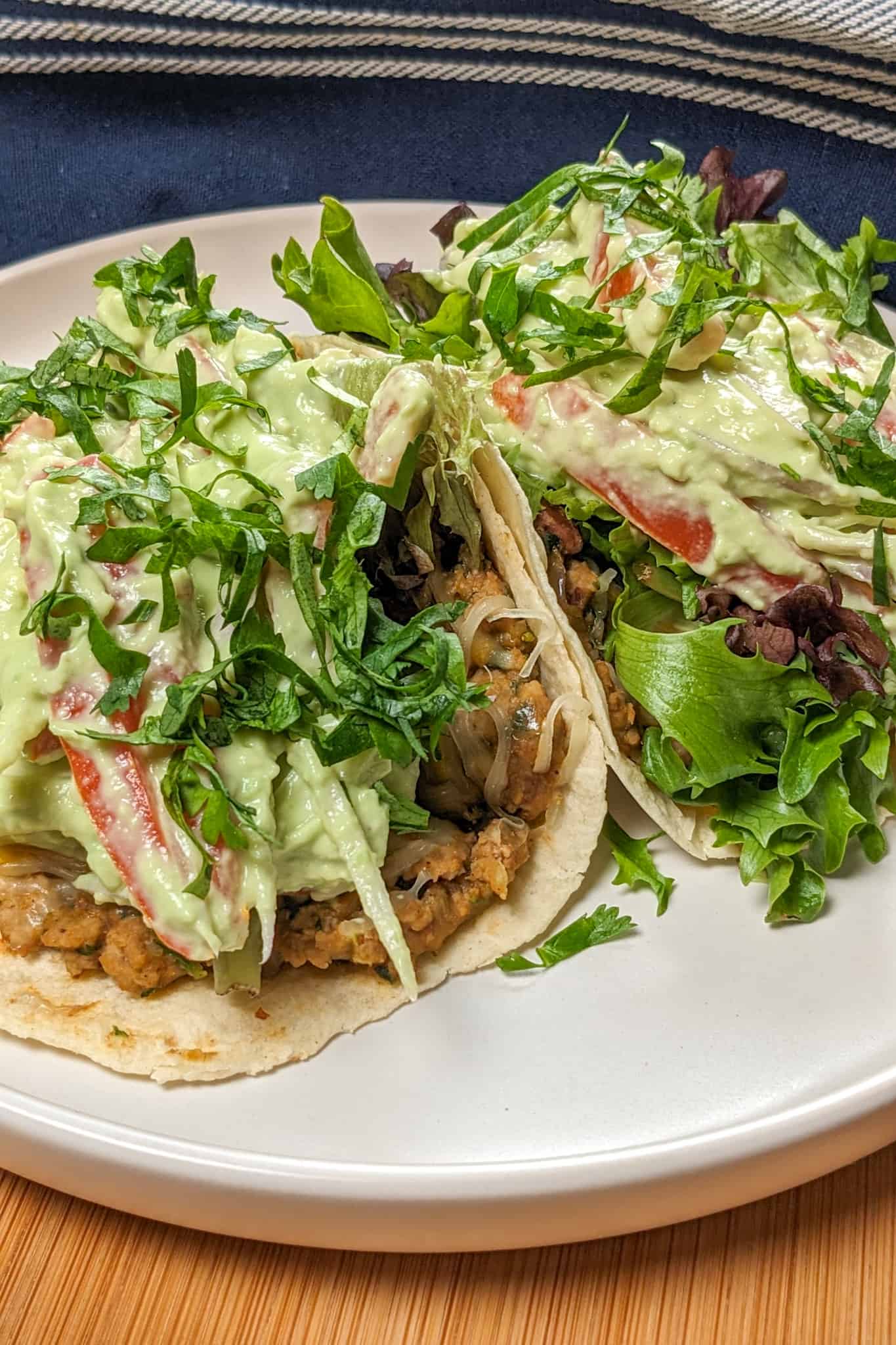 two Salsa Verde Turkey and Beans Tacos with Avocado Cream on a white round plate with shallow sides on a wooden lazy susan with a kitchen towel in the background.