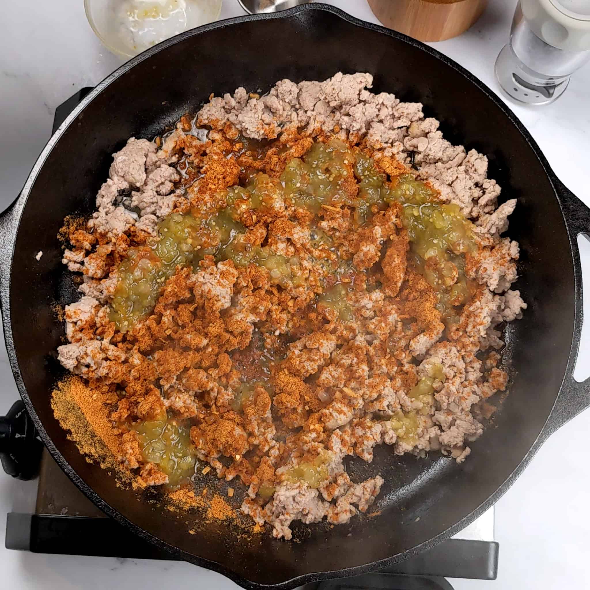 salsa verde adnd taco seasoning covering cooked crumbled ground turkey in a cast iron skillet.