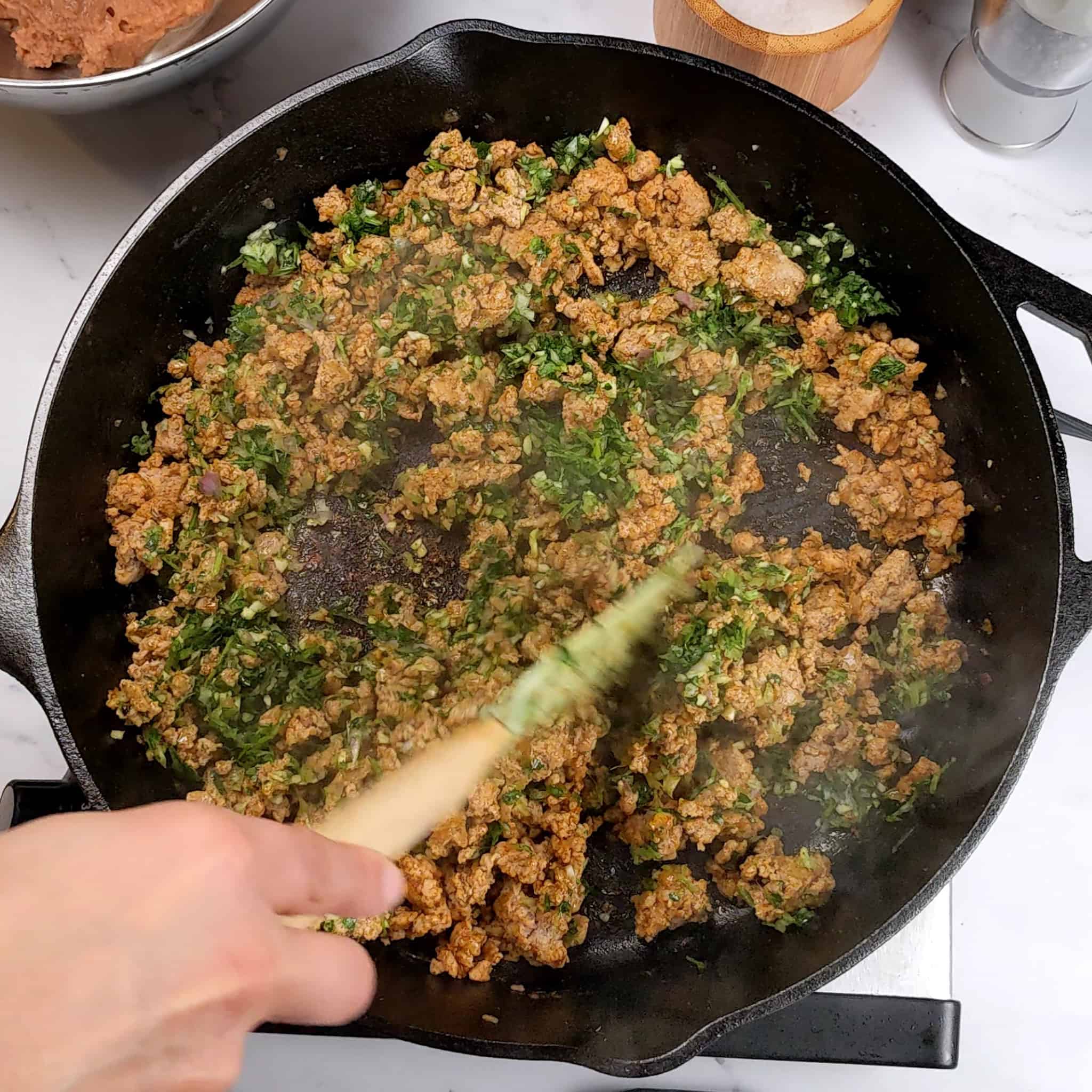 aromatic paste of chopped cilantro, shallots and garlic being mixed into cooked crumbled ground turkey in a cast iron skillet with a silicone spatula.
