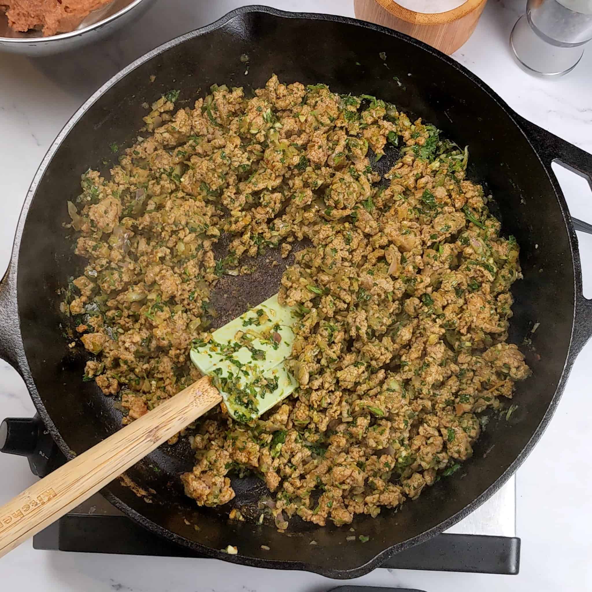 aromatic paste of chopped cilantro, shallots and garlic mixed into cooked crumbled ground turkey in a cast iron skillet with a silicone spatula laying on the side in the skillet.