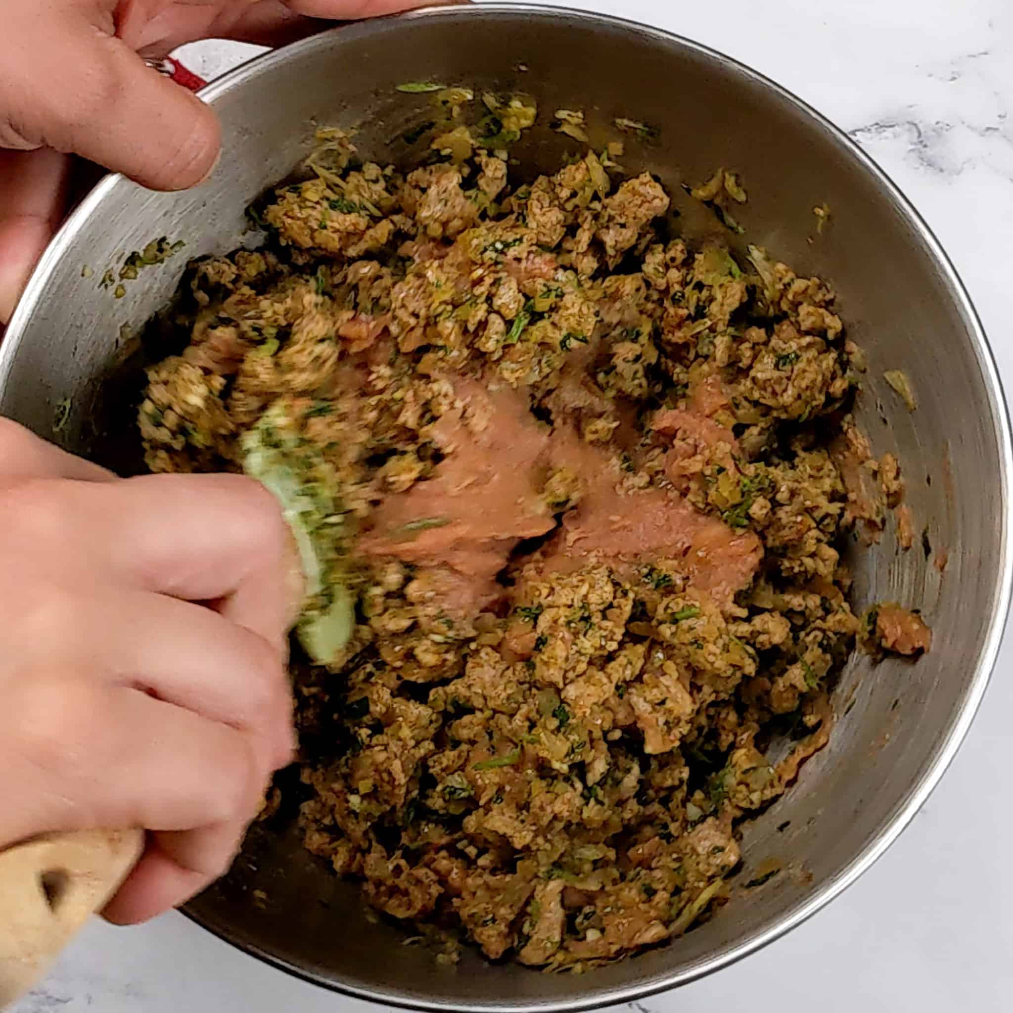 the cooked seasoned ground turkey meat topping the refried beans in a stainless steel mixing bowl being mixed together with a silicon wooden spatula.