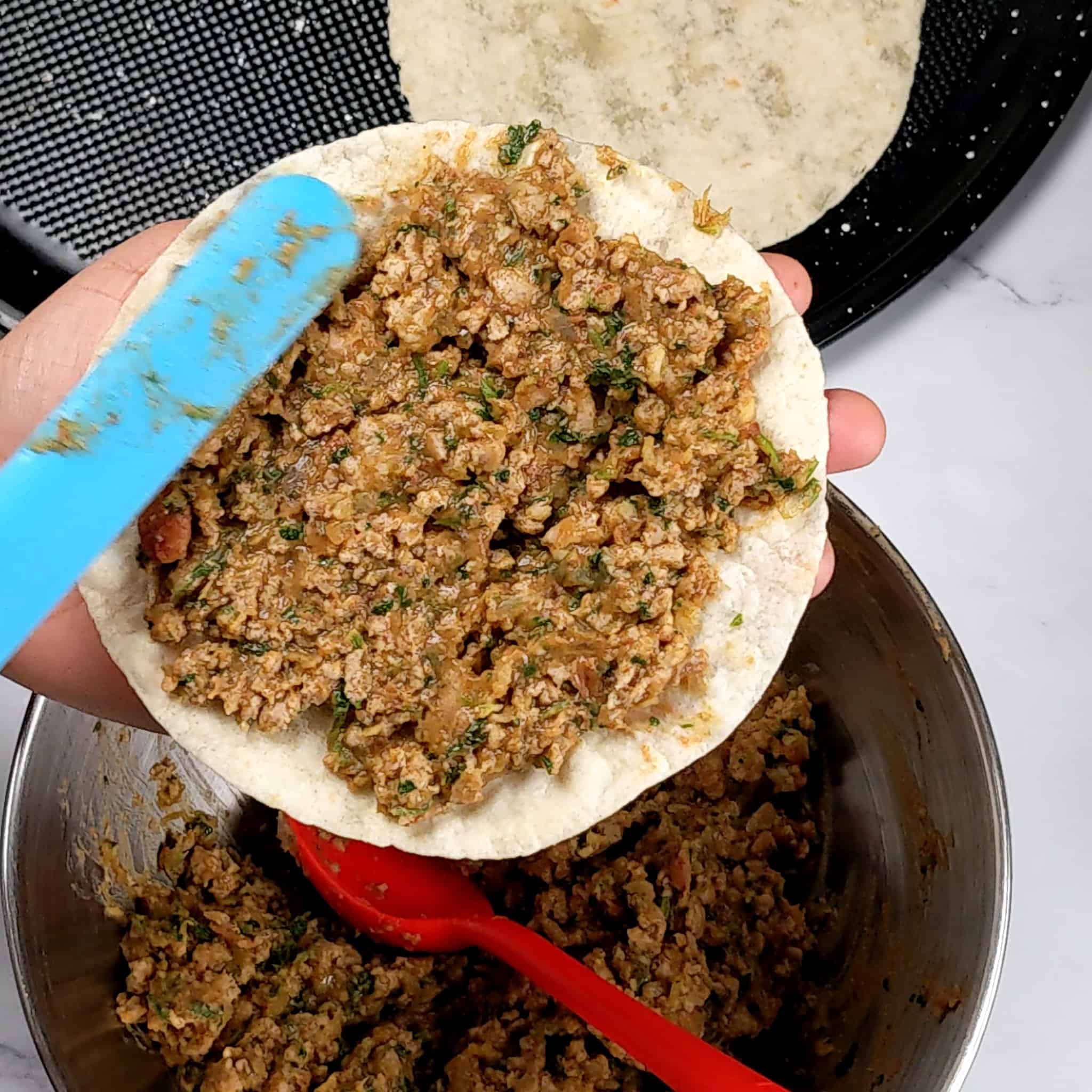 seasoned ground turkey and refried beans mix being spread on a flour tortillas with a long silicone spatula over a bowl of more seasoned meat and a corn tortilla laying in a large pizza pan.