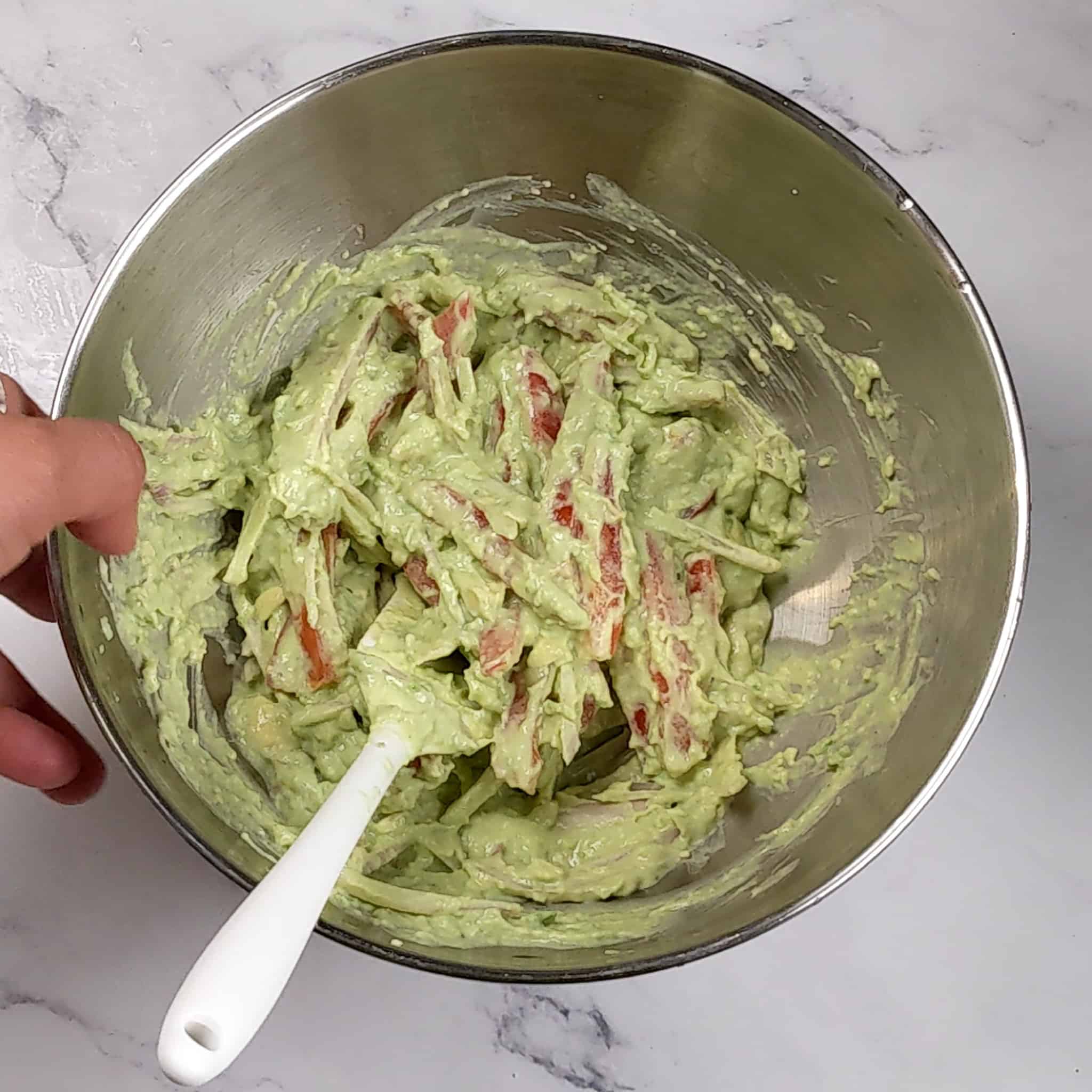avocado cream mixed with thinly sliced shallots and plum tomatoes in a stainless steel mixing bowl with a silicon spatula resting inside.