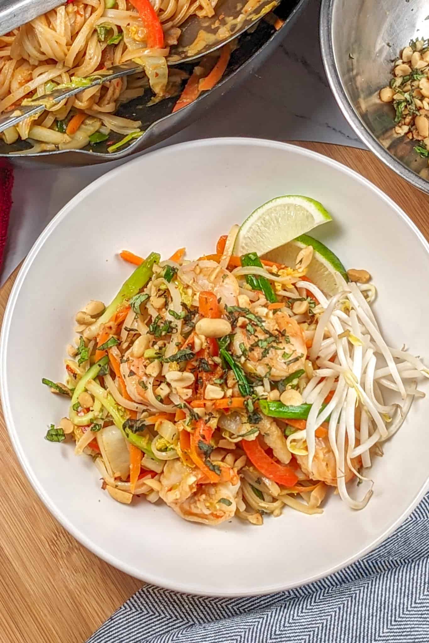 Shrimp and Pork Belly Pad Thai - Spring Roll Style in a wide rim bowl on a wooden lazy susan next to a kitchen towel, a wok with the rest of the noodles and a bowl of toasted peanuts tossed in herbs.