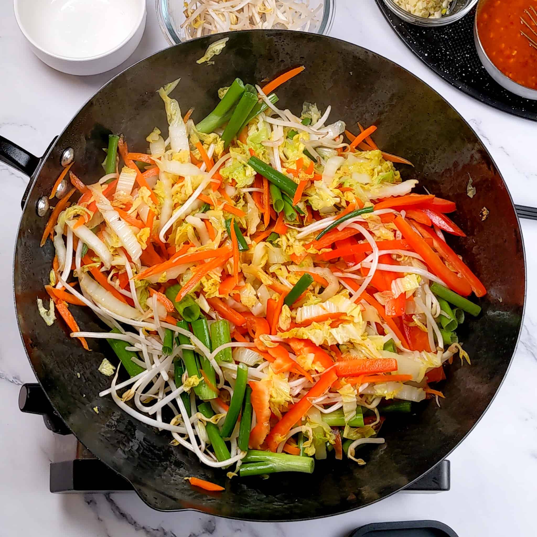 raw red bell pepper, scallions, and bean sprouts, added to the nappa cabbage and carrots in a wok.