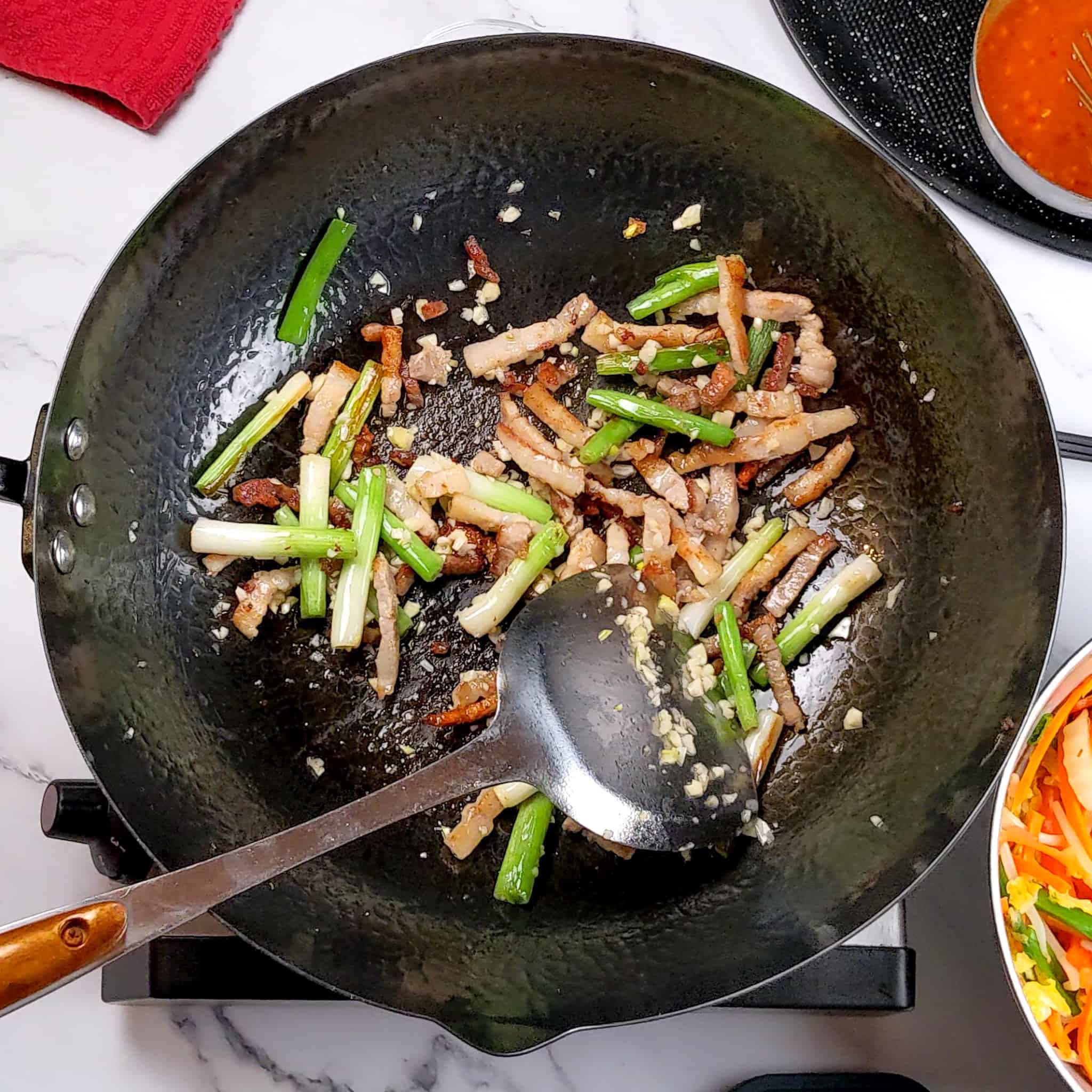 browned chopped garlic, 1-inch scallions and thin strips of pork belly in a wok, with a wok spatula laying on the inside.
