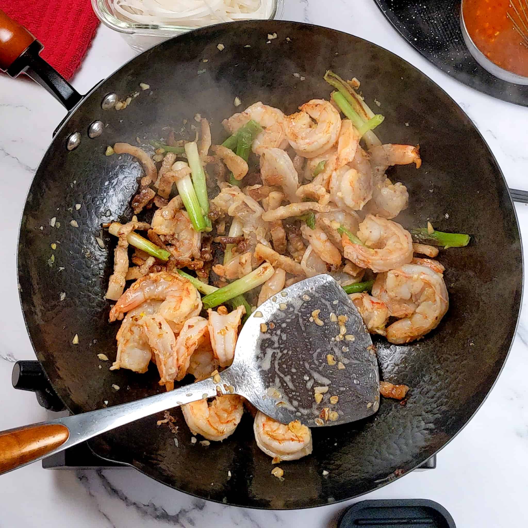 half-way cooked shrimp with browned chopped garlic, 1-inch scallions and thin strips of pork belly in a wok, with a wok spatula laying on the inside.