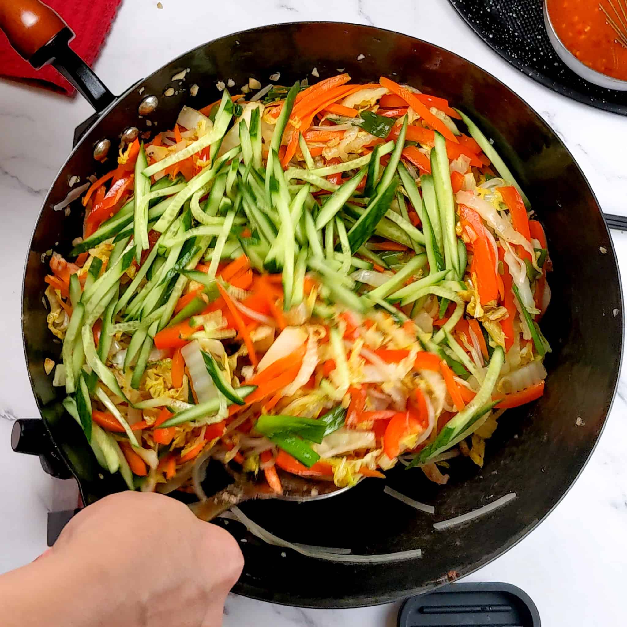 stir fried vegetables and cucumbers added to the wok and are being mixed together with a wok spatula.