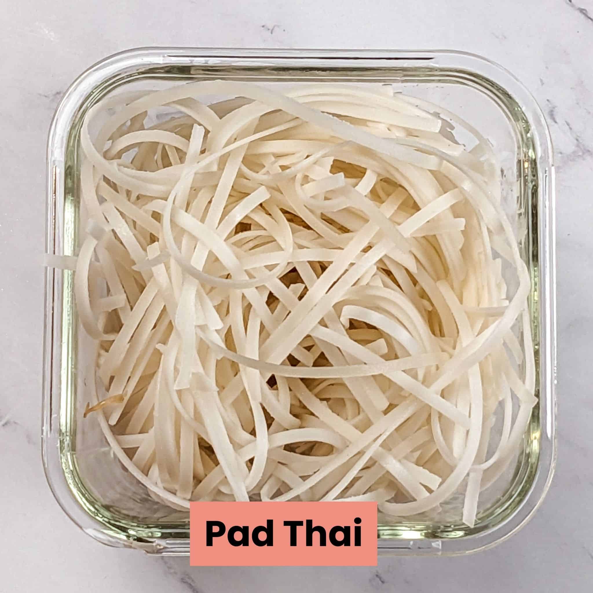 soaked and drained pad thai noodles in a deep square glass container.