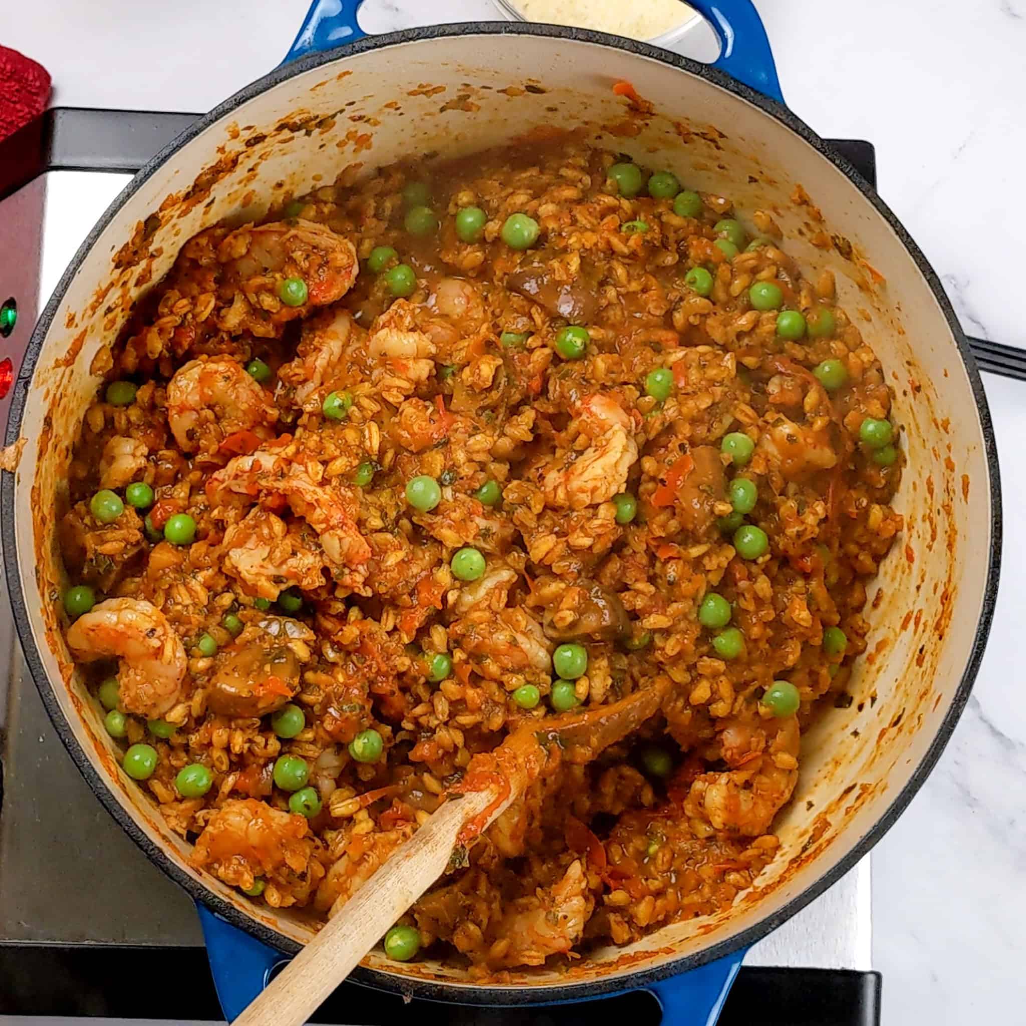 cooked Spicy Shrimp Farro Risotto with Mushrooms and Peas in a lodge enameled dutch oven with a wooden spoon.