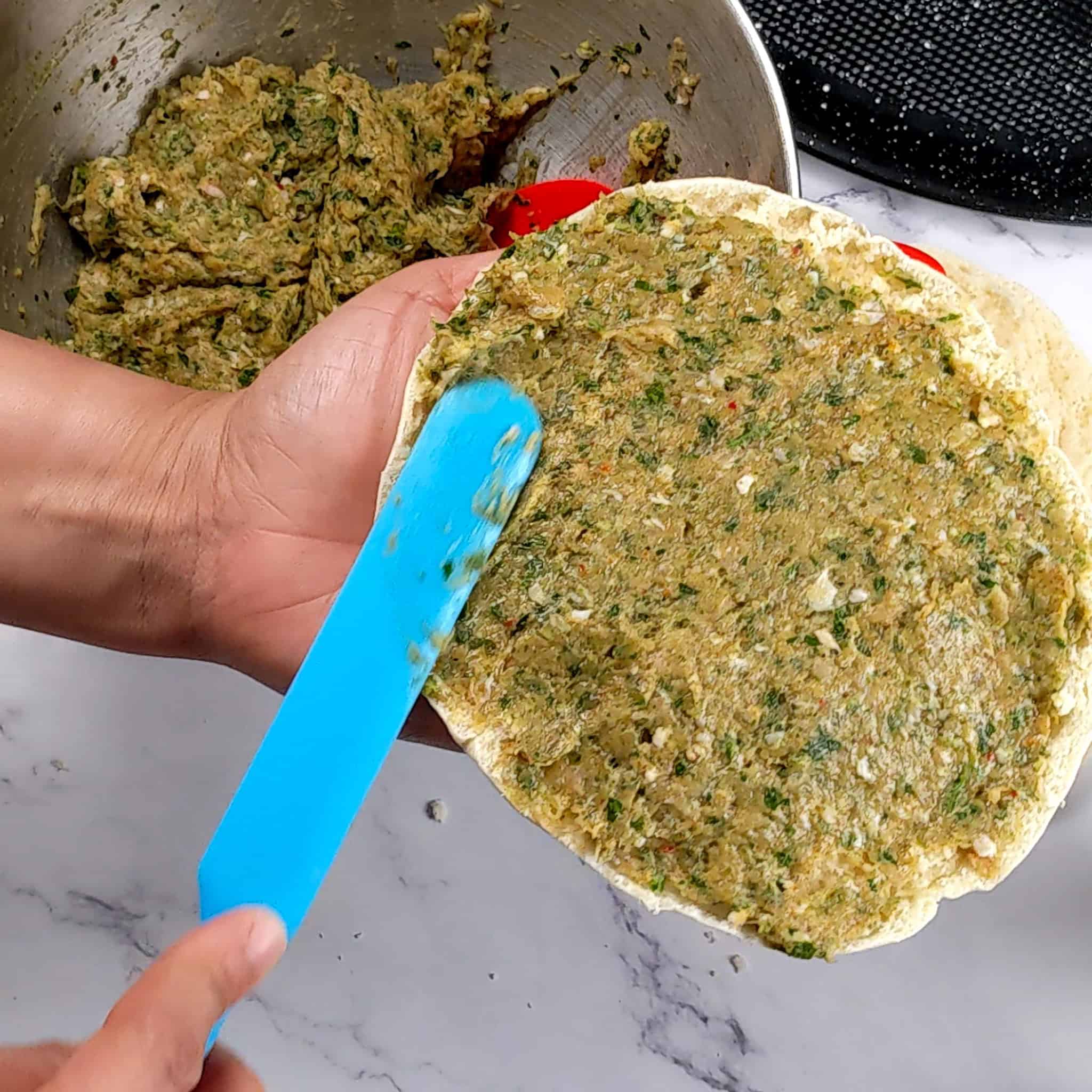 the spicy zhug ground chicken mixture being spread on one half of a pita round with a long silicone spatula.
