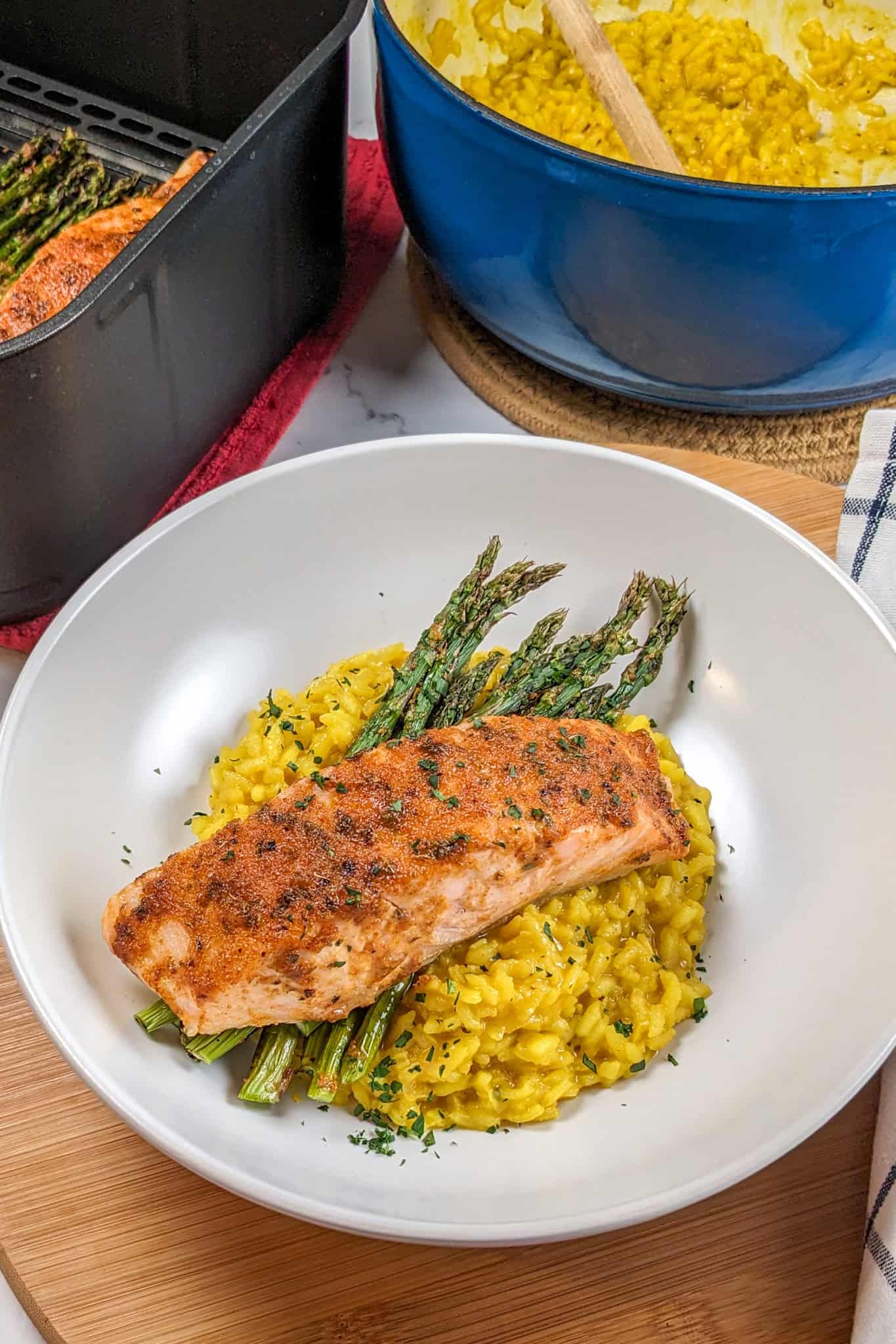 Air Fryer Habanero Adobo Salmon and asparagus with Yellow Risotto on a round wide rim bowl next to a kitchen towel, the cosori air fryer basket and lodge enamel dutch oven with the yellow risotto.