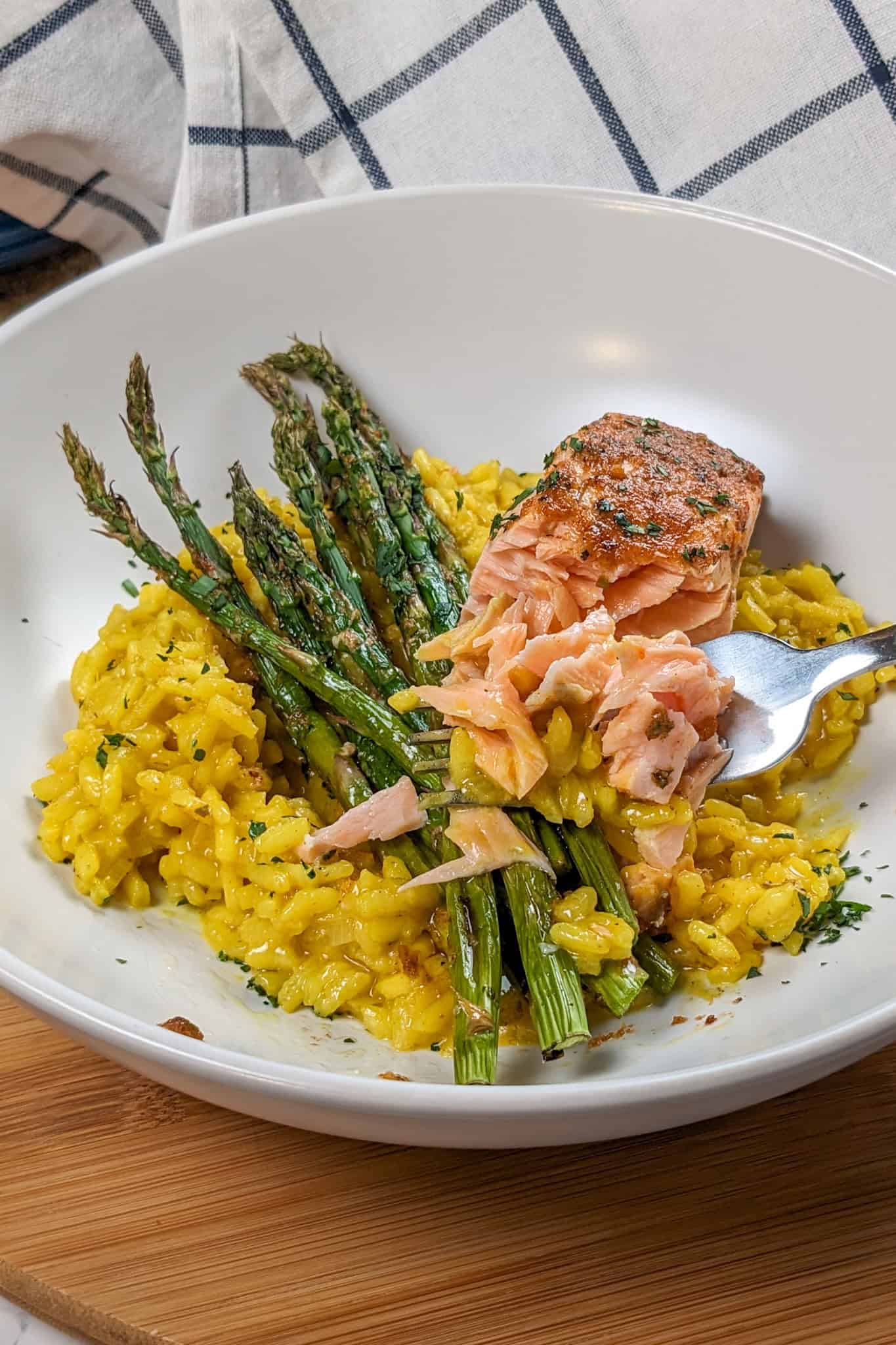 top view of the Best Air Fryer Habanero Adobo Salmon and Yellow Risotto with asparagus on a round wide rim bowl next to a kitchen towel with a fork with some of the risotto and salmon on it.