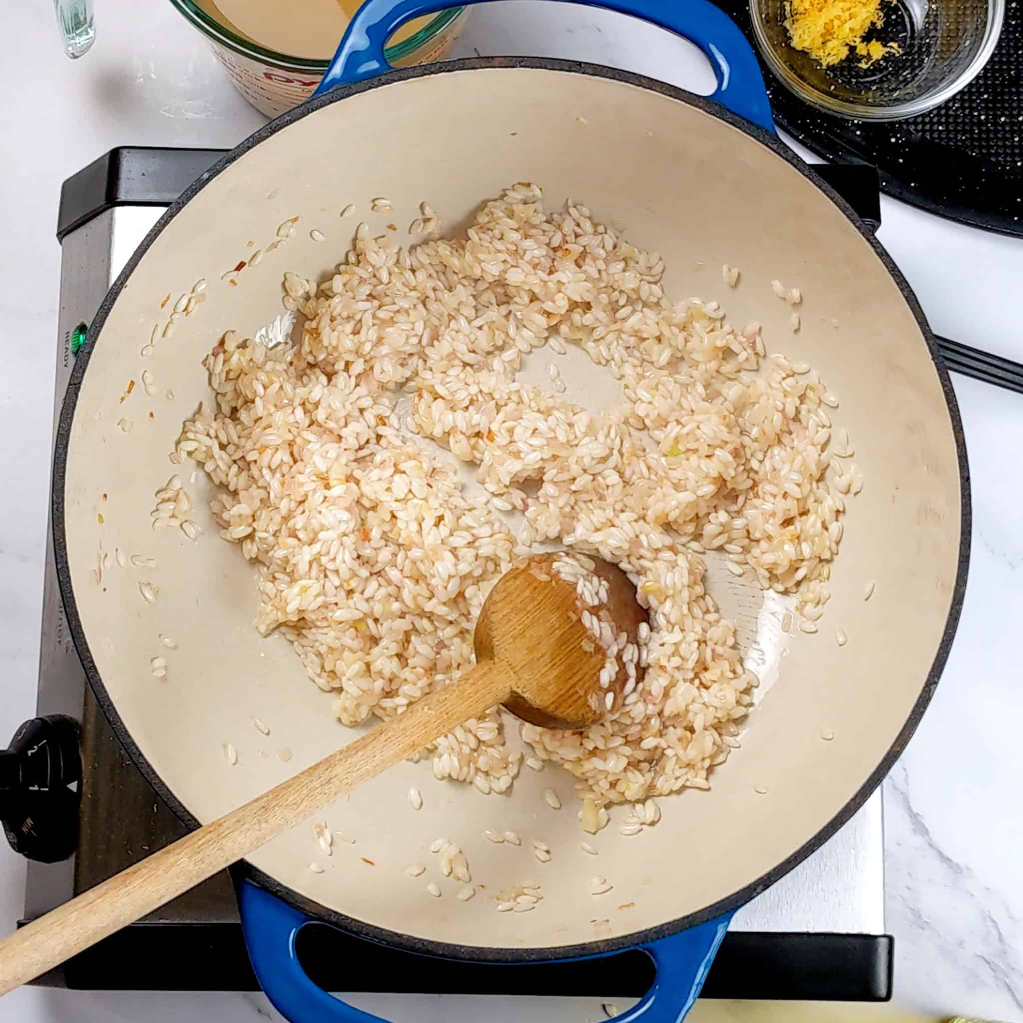 white wine soaked arborio rice in a lodge enamel dutch oven being stirred with a wooden spoon.
