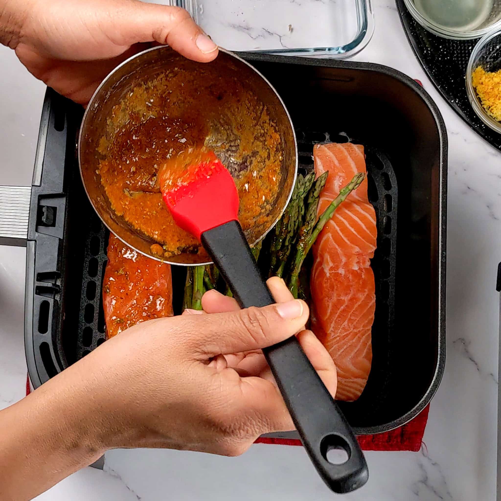 wet adobo seasoning in a small stainless steel bowl with a silicone brush over the cosori basket wtih raw salmon and asparagus.