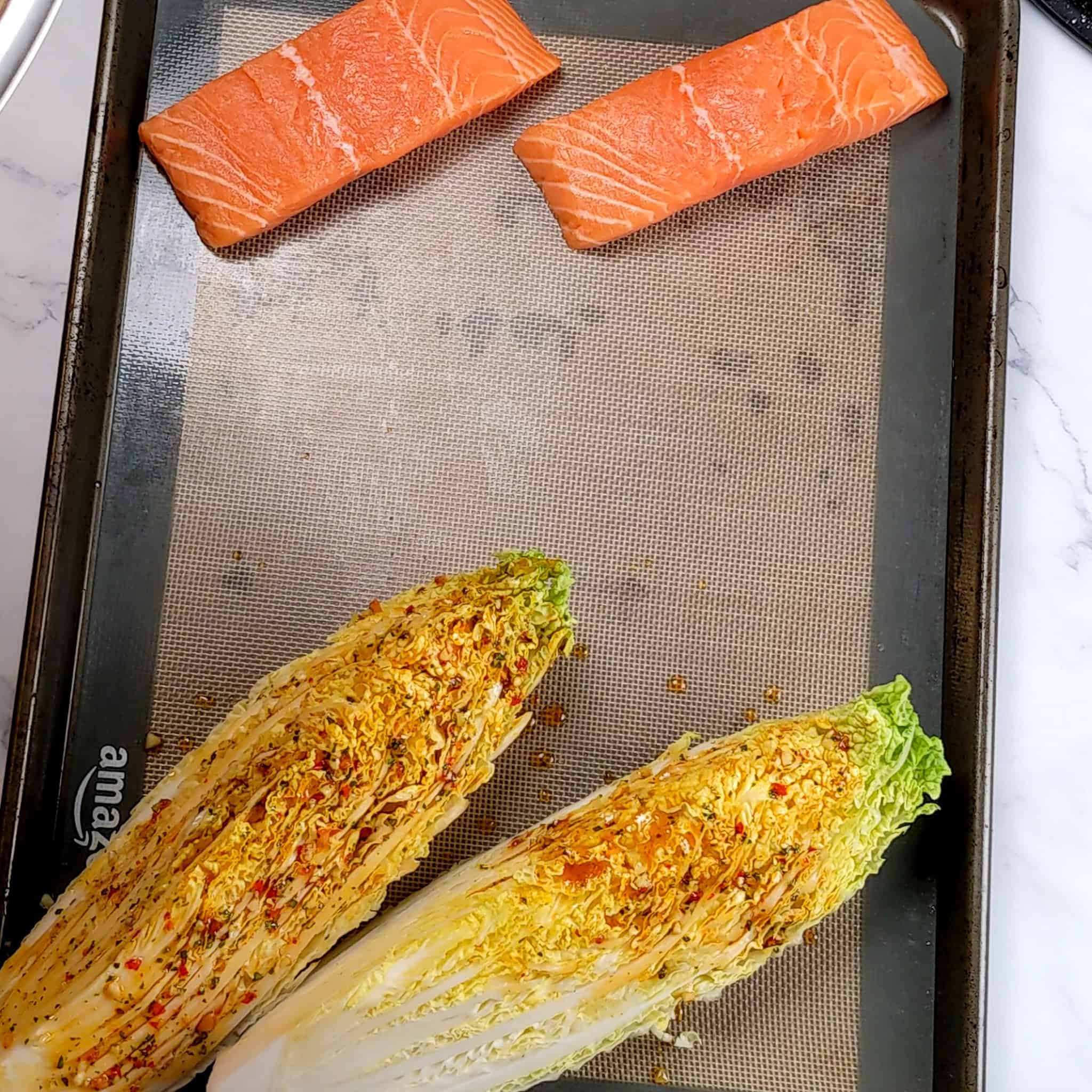 a sheet pan lined with a silicon liner with salmon filets on one side lined vertically on a diagonal and napa cabbage wedges on the other far side in the same manner with some of the wet seasoning on it.