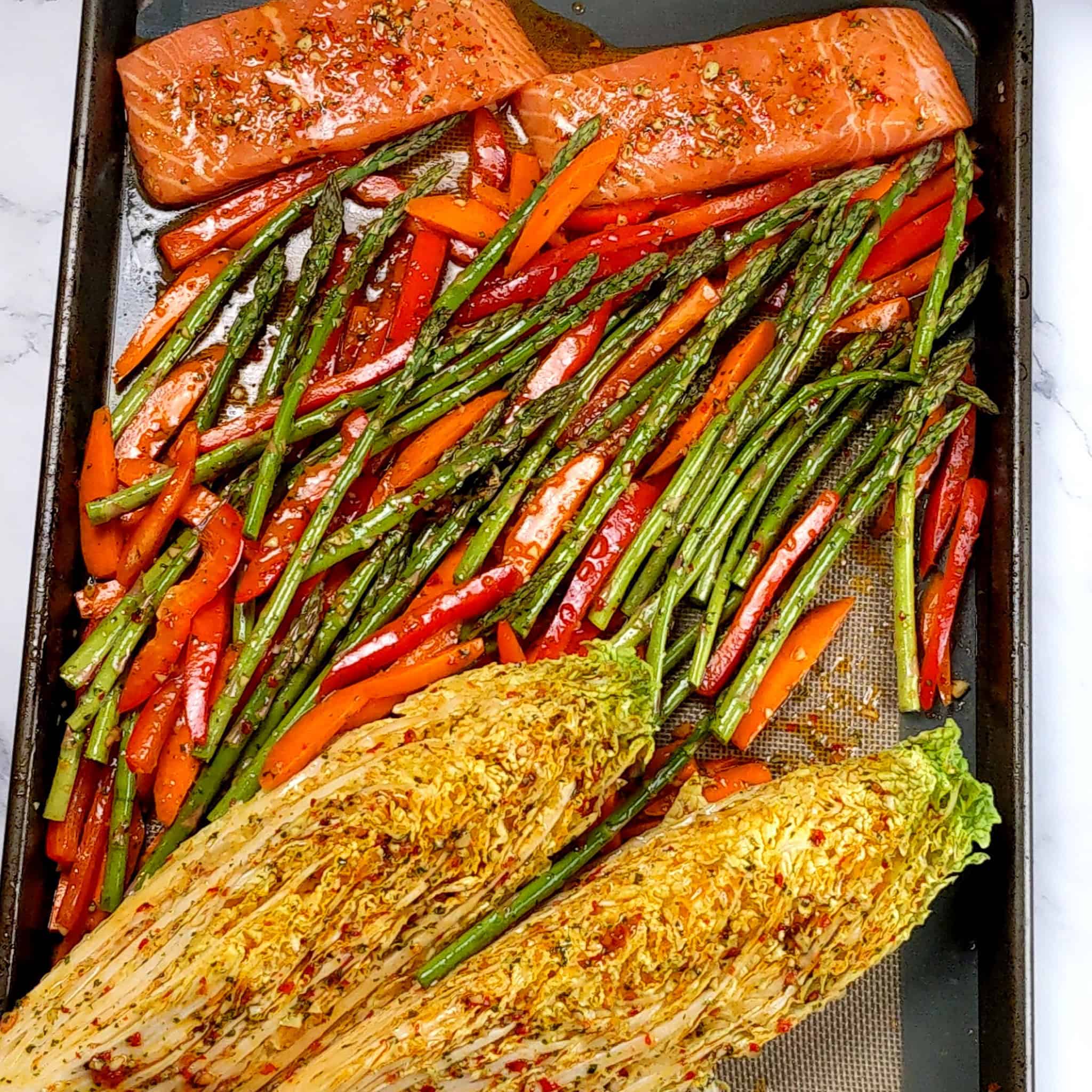 the  chili lime salmon, vegetables and napa cabbage arranged in lines on the silicone lined sheet pan dressed in the wet seasoning.