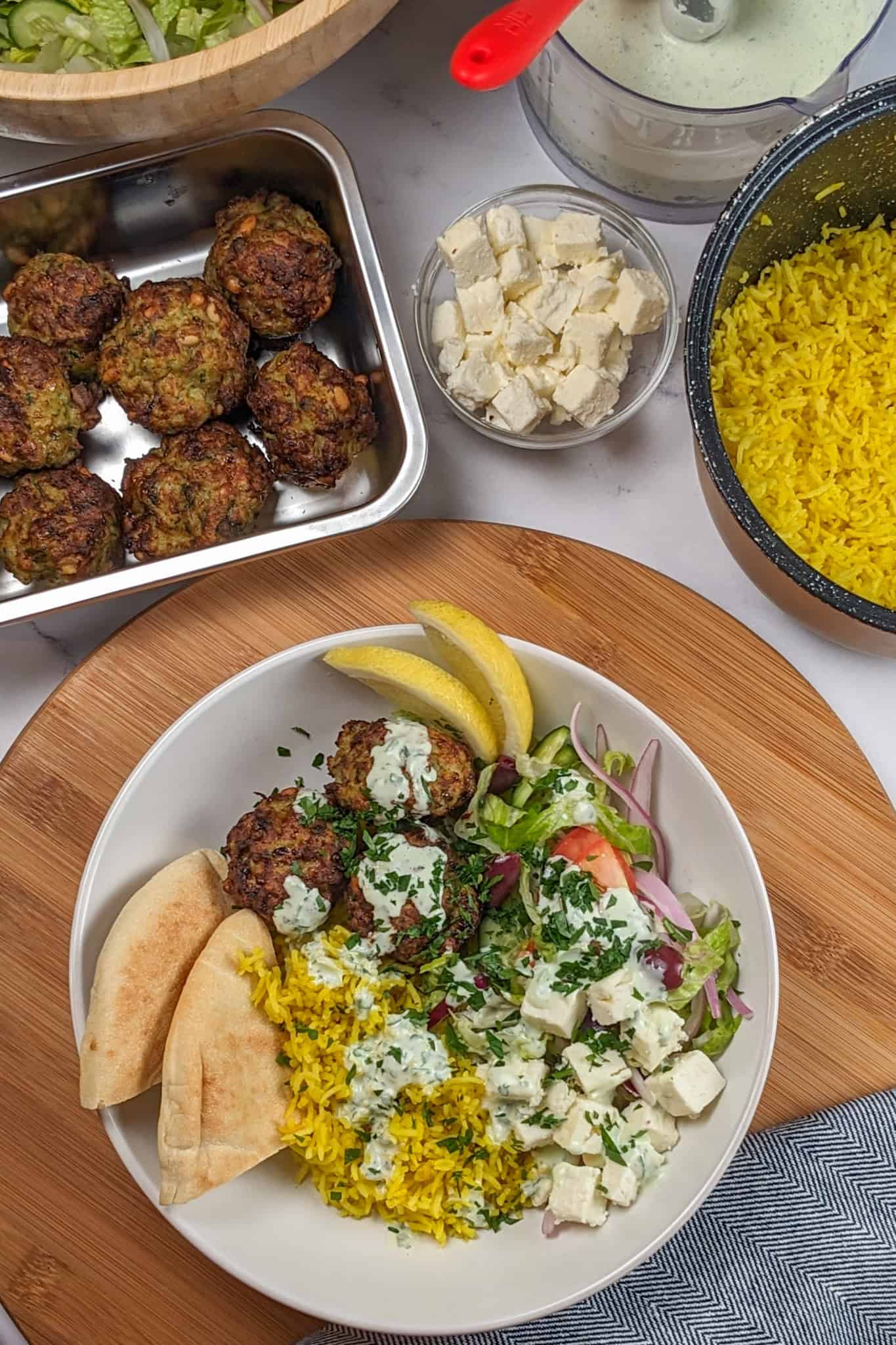 top view of the Spicy Greek Chicken Meatball Rice Bowl in a wide rim bowl garnished with chopped parsley, Greek yogurt sauce, two pita triangles and lemon wedges; surrounded by a stainless steel rectangle container of cooked meatballs, wooden bowl with salad, glass round dish with large diced feta cheese, the food processor container with the yogurt sauce and the rice cooker canister with the spiced yellow basmati rice.