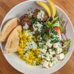 The Spicy Greek Chicken Meatball Rice Bowl in a wide rim bowl drizzled with yogurt sauce and garnished with chopped parsley, pita triangles and lemon wedges.