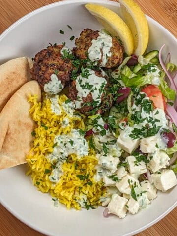 The Spicy Greek Chicken Meatball Rice Bowl in a wide rim bowl drizzled with yogurt sauce and garnished with chopped parsley, pita triangles and lemon wedges.