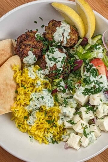 The Spicy Greek Chicken Meatball Rice Bowl in a wide rim bowl drizzled with yogurt sauce and garnished with chopped parsley, pita triangles and lemon wedges.