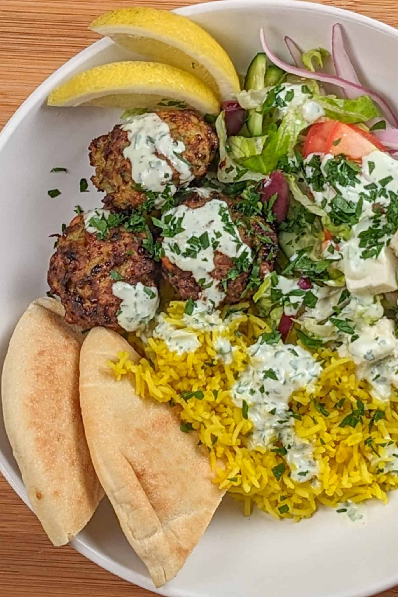 The Spicy Greek Chicken Meatball Rice Bowl in a wide rim bowl drizzled with yogurt sauce and garnished with chopped parsley, pita triangles and lemon wedges.