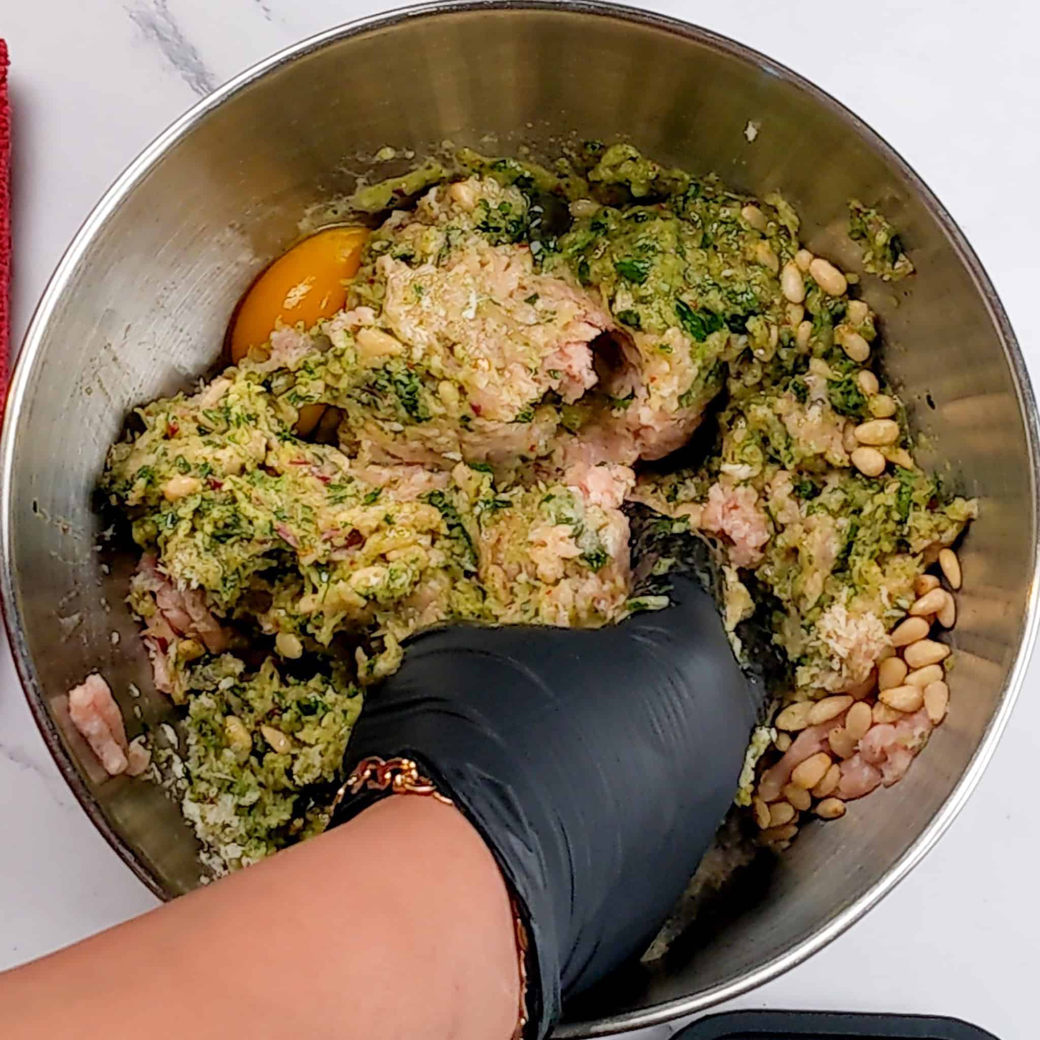 a stainless steel bowl with the meatball mixture being combined with a gloved hand.