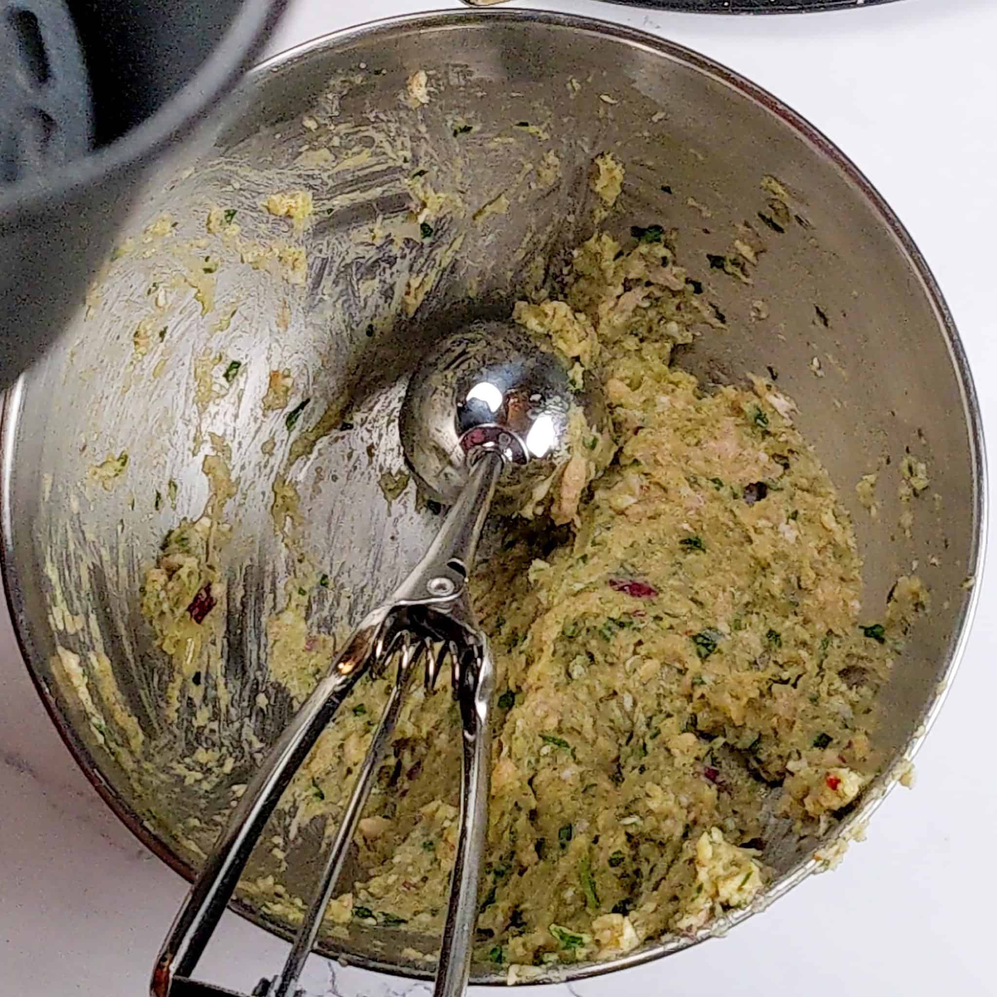 a medium cookie scoop resting in the stainless steel bowl of the raw meatball mixture.