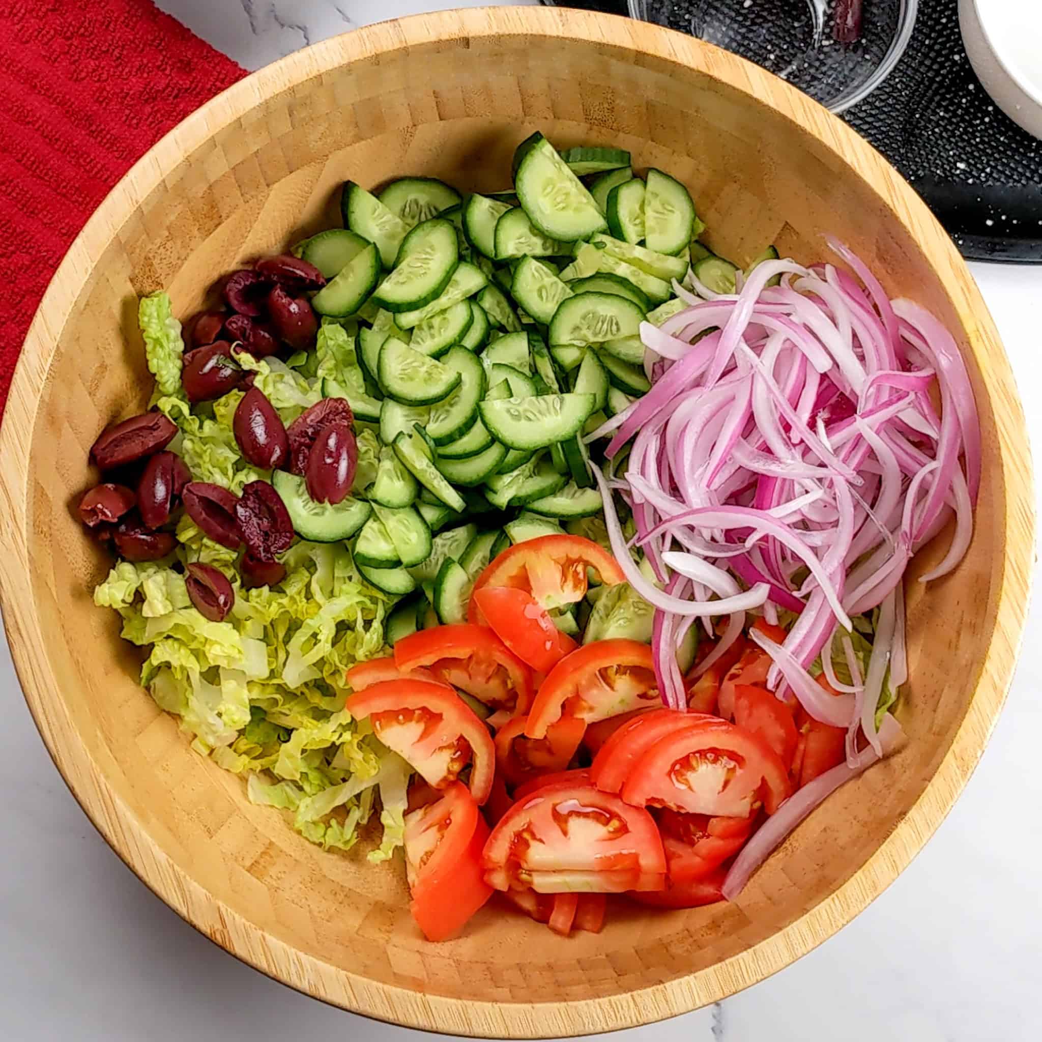 a wooden bowl with shredded romaine topped with sliced cucumber, red onions, tomatoes and kalamata olives.