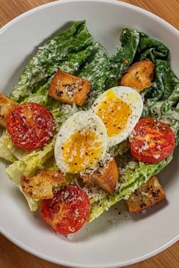 Caesar Salad with soft-boiled Egg, roasted campari Tomatoes and Chili-Infused Croutons garnished with freshly grated parmesan cheese and cracked black pepper in a wide rim bowl.