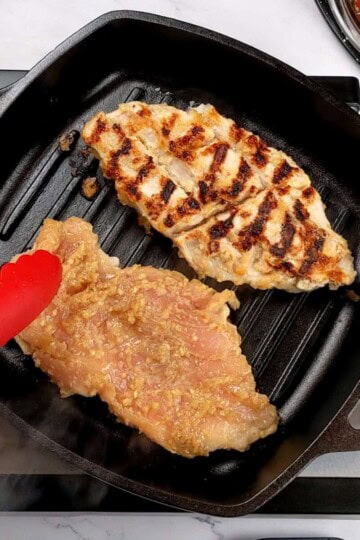 two grilled lemongrass chicken breast with grill marks on one and another about to be flipped with a kitchenaid silicone spatula in a grill pan.