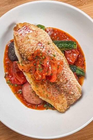 Pan seared snapper toped with stewed onion and red bell pepper garnish on a bed of roasted baby vegetables on spicy creole sauce in a wide rim round bowl.