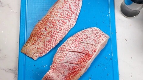 slitted salt and pepper seasoned red snapper filets on a color coded plastic cutting board for raw fish .